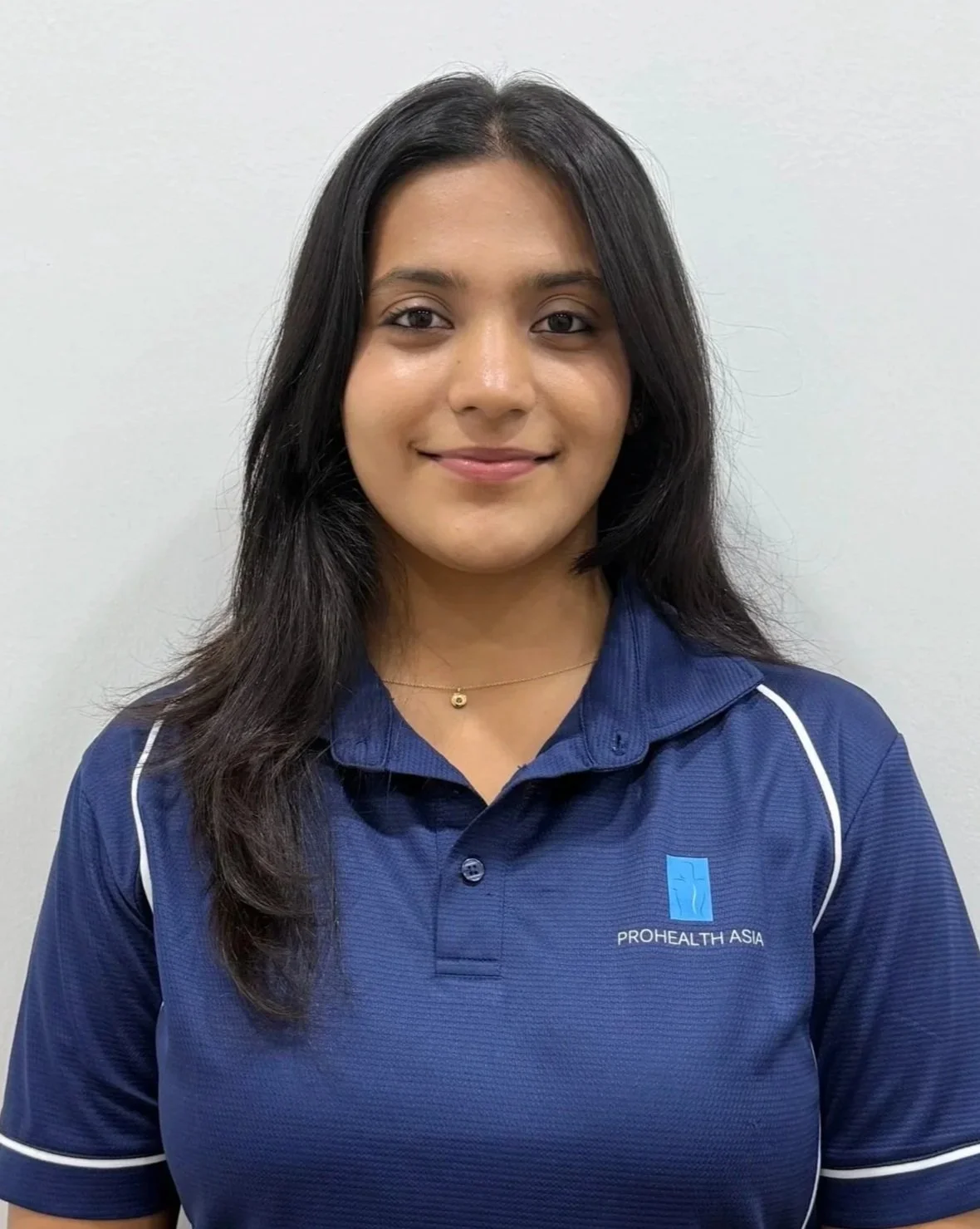 A woman with long dark hair wearing a blue ProHealth Asia shirt, smiling, standing against a plain white wall.
