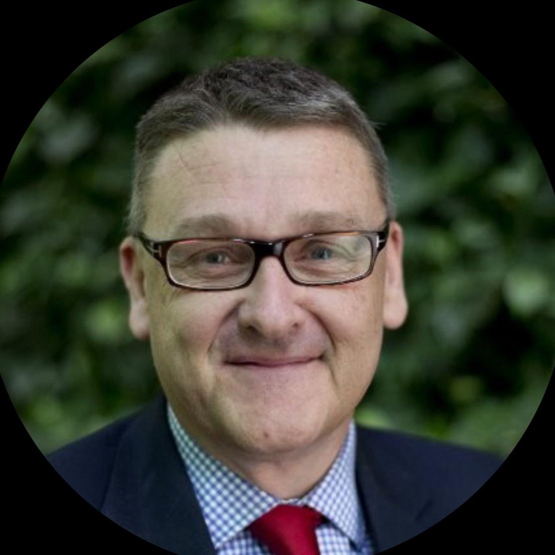 Close-up of a smiling man wearing glasses, a suit, a checkered shirt, and a red tie, with a blurred green outdoor background.