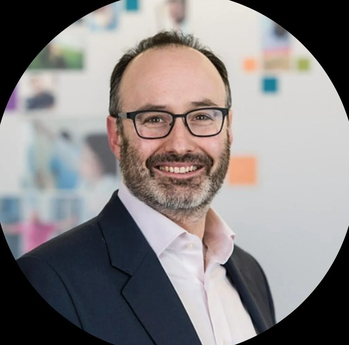 A smiling man with glasses, a beard, and short hair, dressed in a suit and pink shirt, standing in a modern office with colorful sticky notes on the wall behind him.