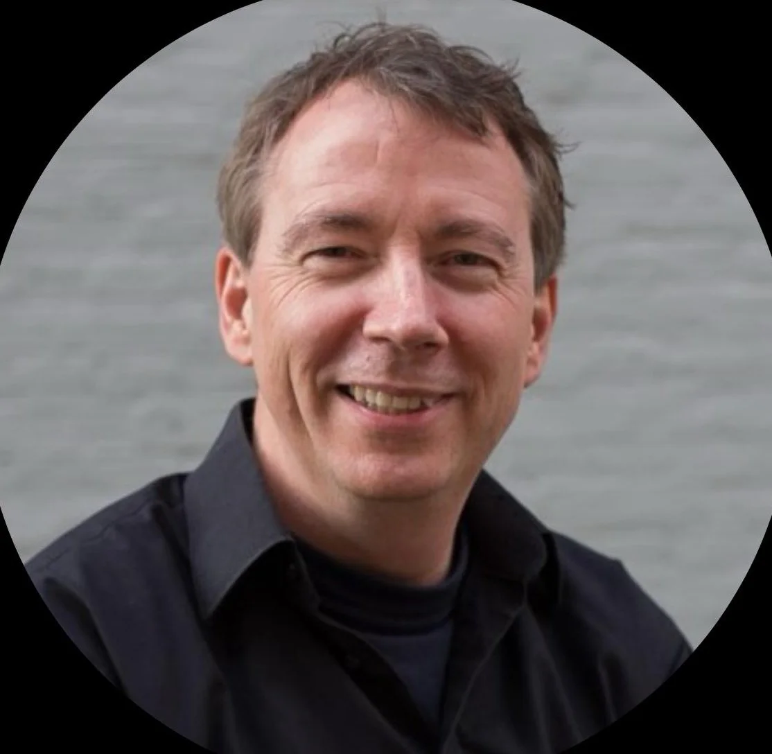 A smiling man with short brown hair wearing a black shirt outdoors near water.