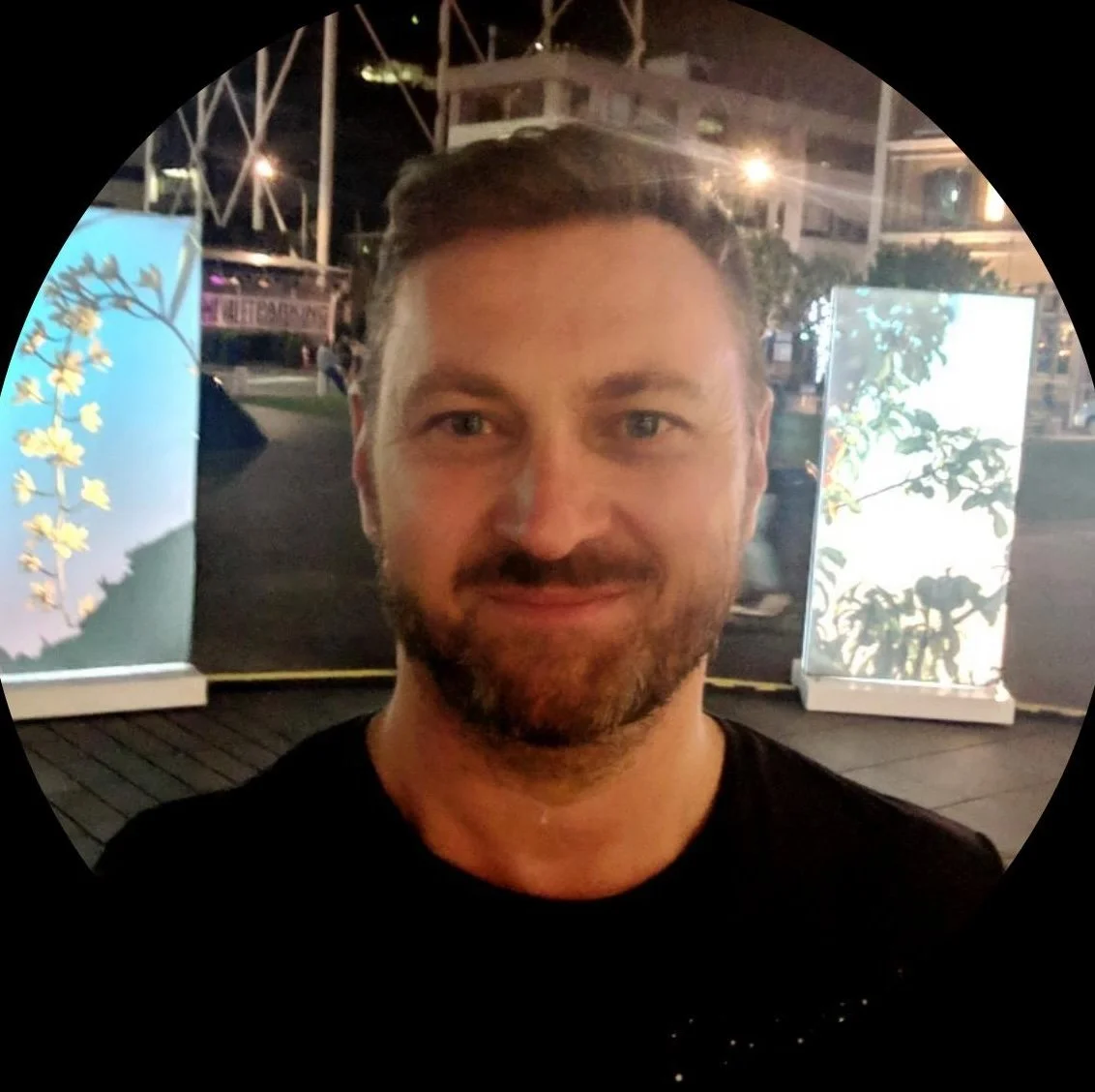 A man with short brown hair, light beard, and blue eyes taking a selfie at night. In the background, illuminated display boards with botanical illustrations are visible.