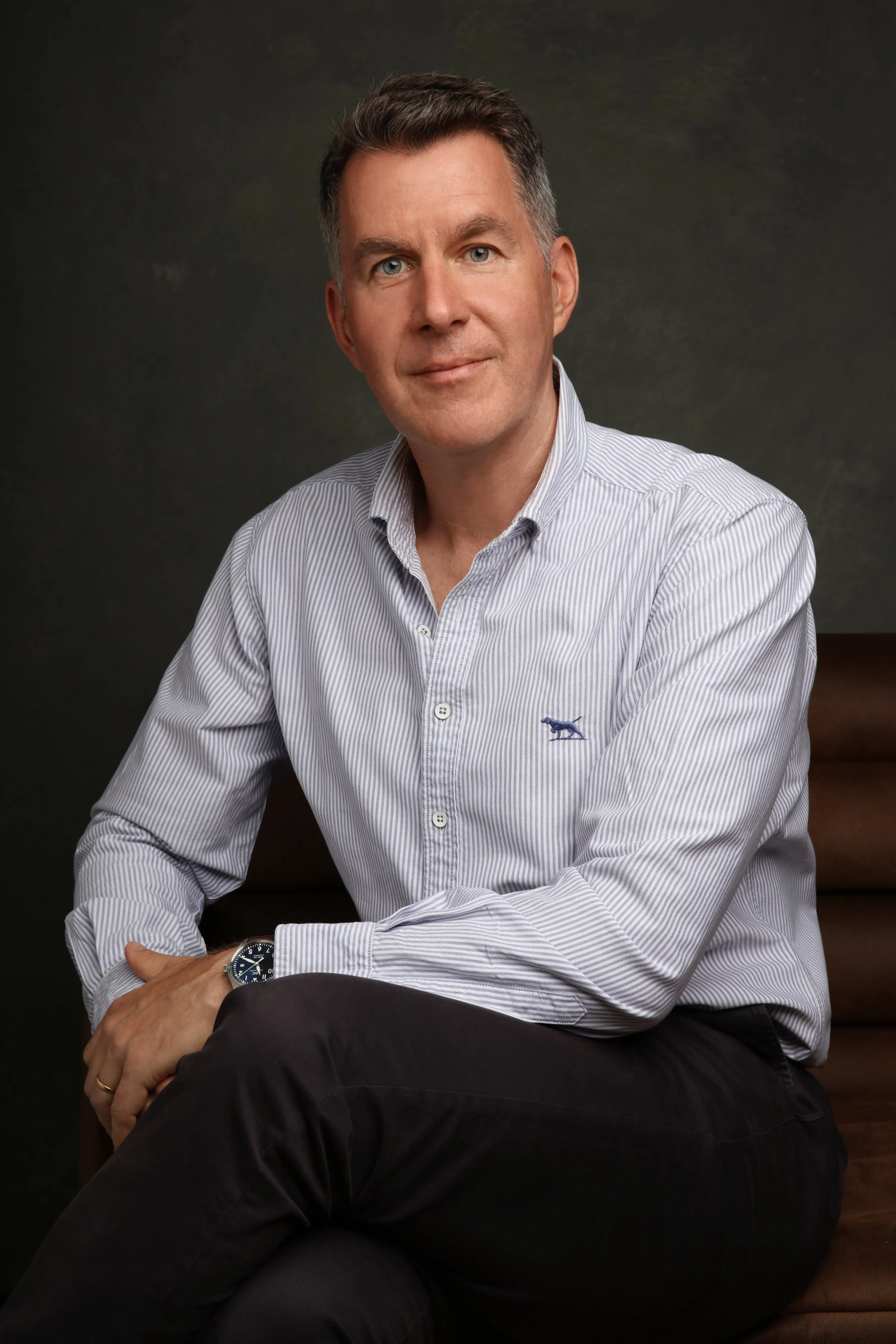 A man with short dark hair and blue eyes sitting on a brown chair, wearing a light blue striped button-up shirt and black pants, with a watch on his left wrist, against a dark background.