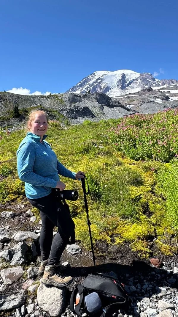Cheryl Scott standing on a mountain trail near Mount Rainier, camera in hand and trekking pole planted in the ground, surrounded by wildflowers and moss, with the snow-capped peak in the background.