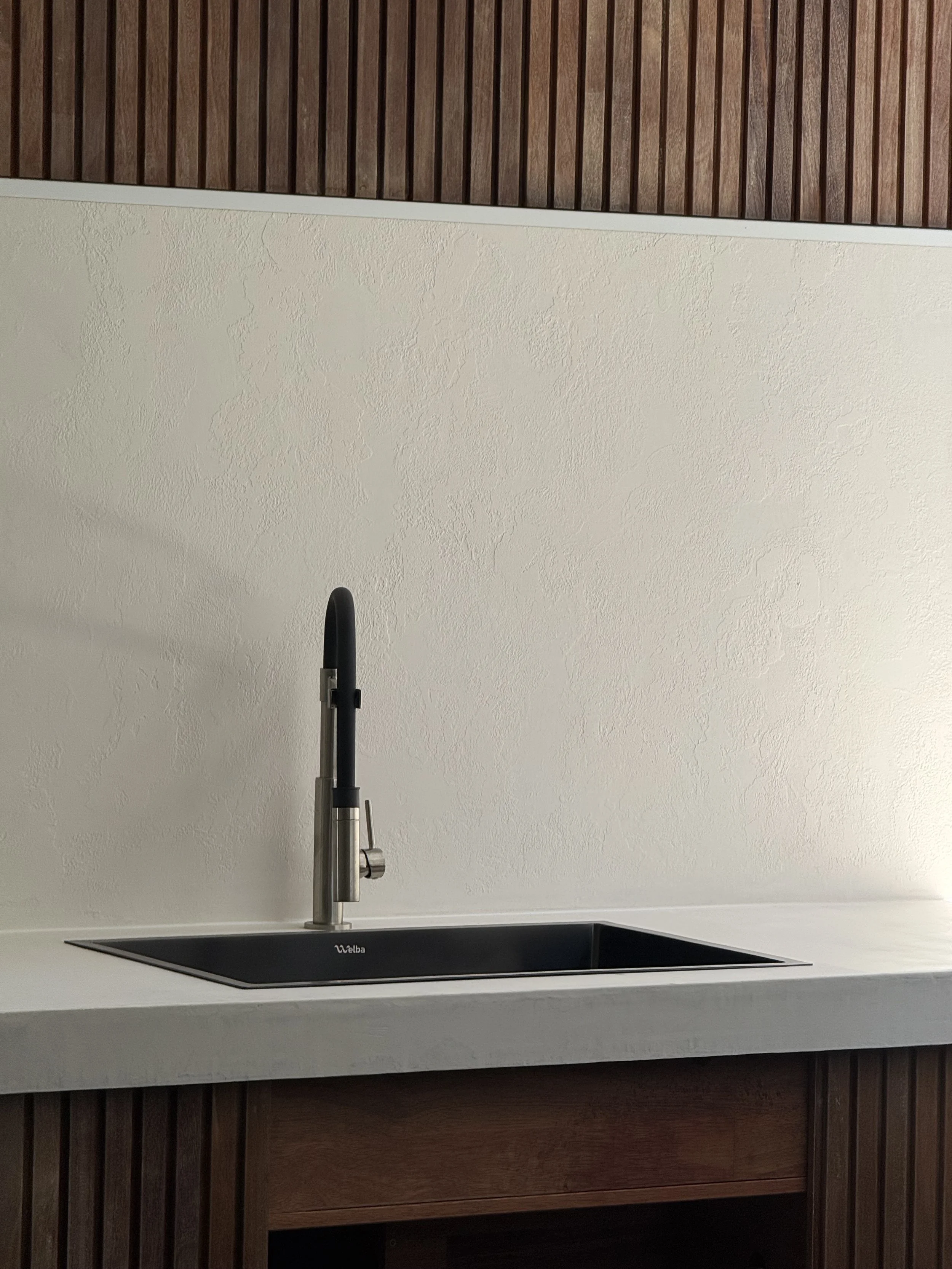 Kitchen sink area with a black undermount sink, stainless steel faucet, white countertop, beige textured wall, and brown wooden cabinetry above and below.