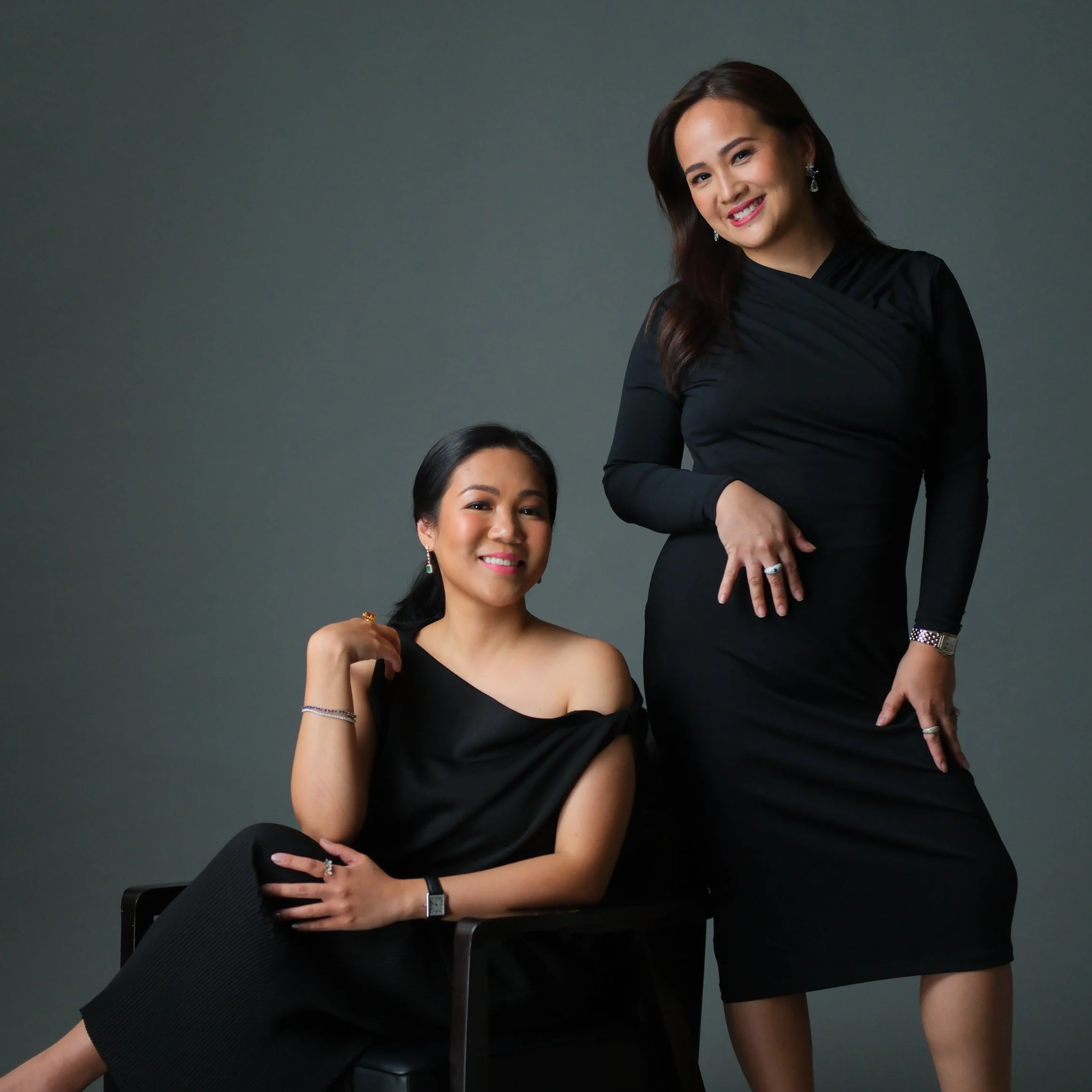Two women dressed in black, one sitting on a chair with a relaxed pose and big smile, the other standing slightly behind her, wearing earrings and rings, against a plain gray background.