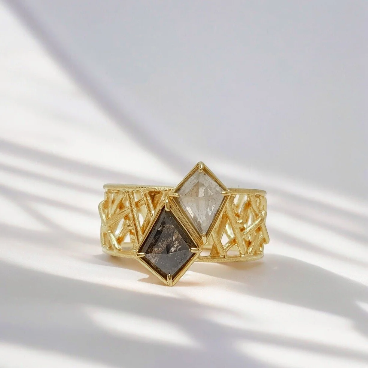 Gold ring with three rectangular gemstones in clear, dark, and smoky colors, set in a geometric design, with shadows cast on a white surface.