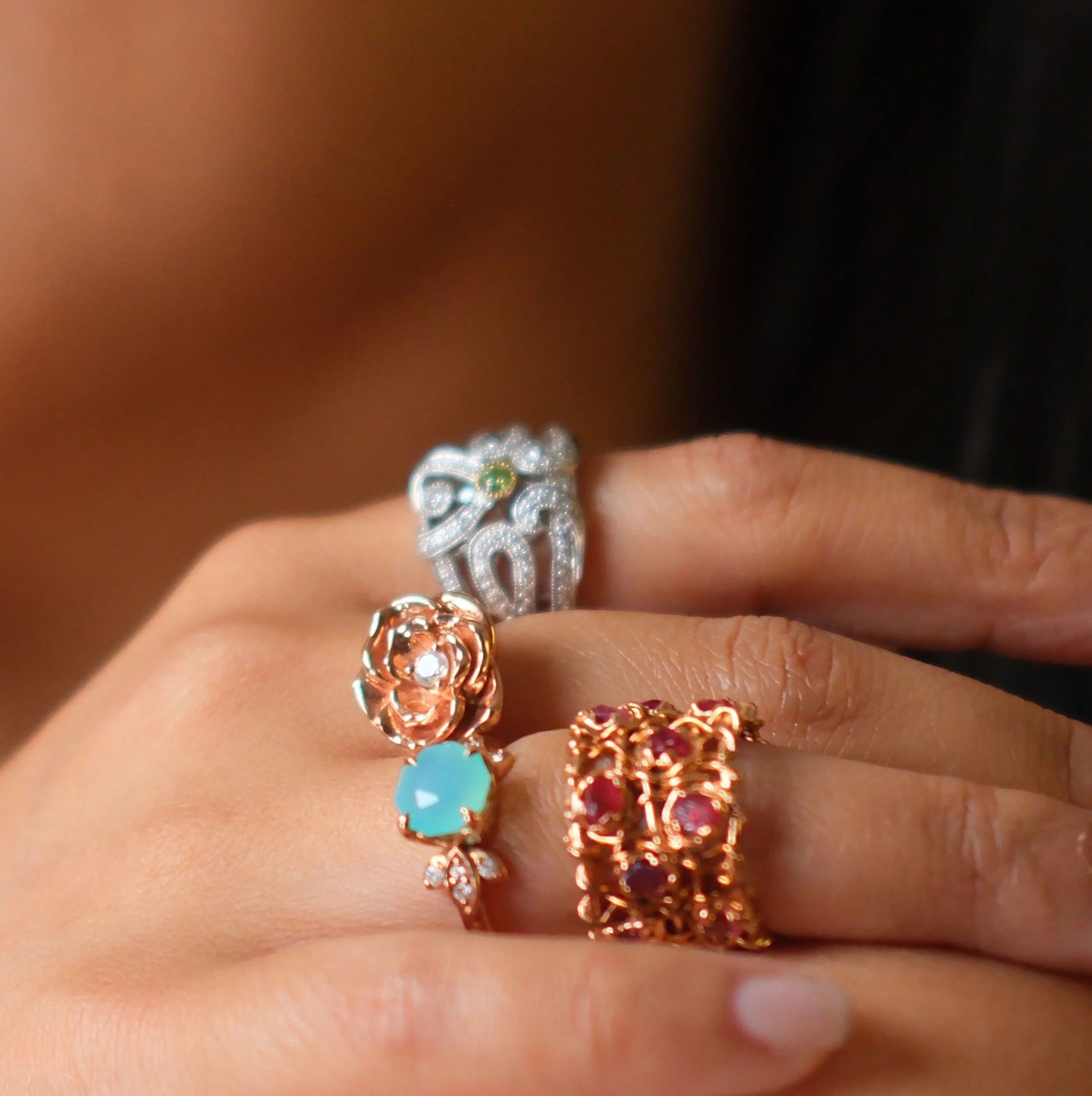 Close-up of a hand wearing three rings, each with intricate designs and gemstones, against a dark background.