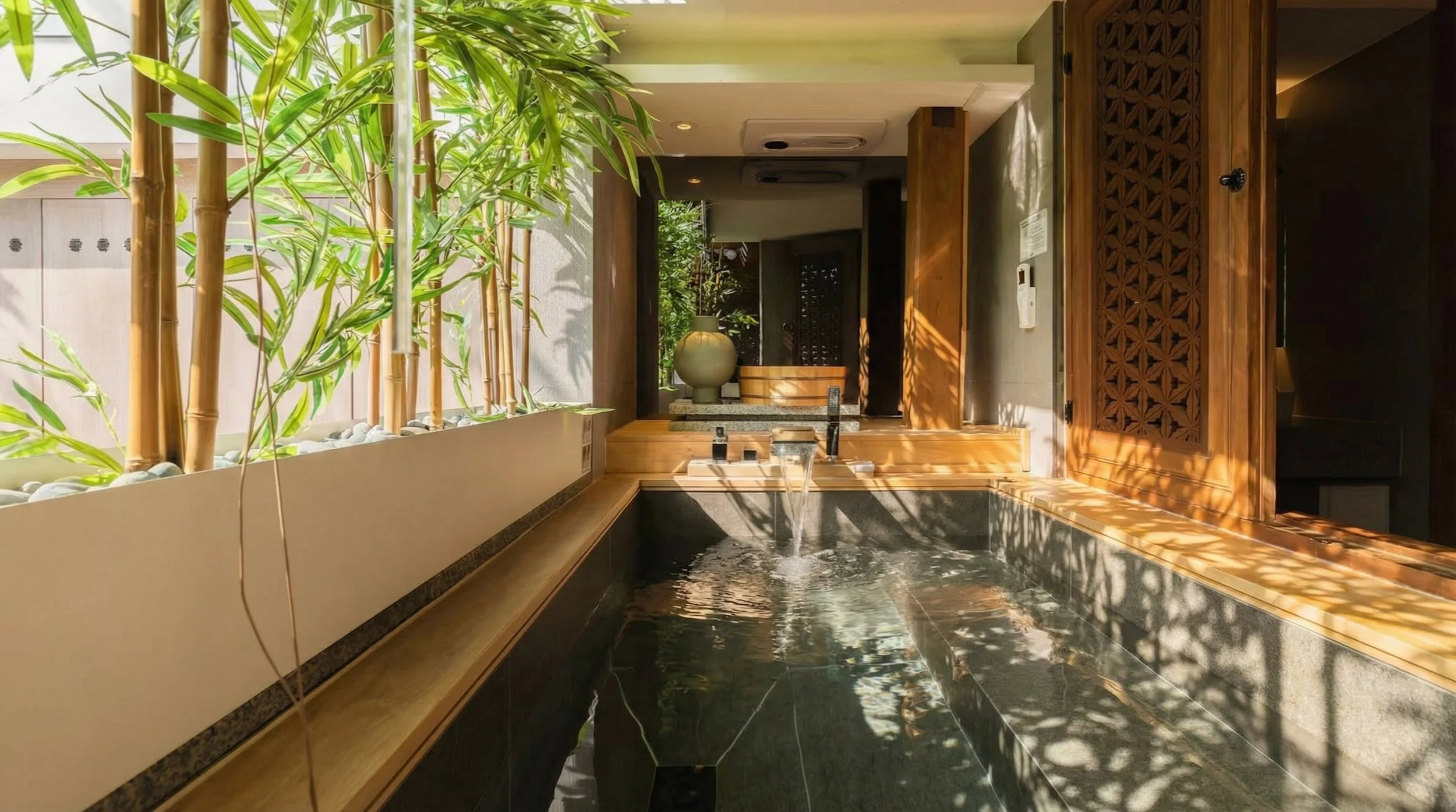 Sunlit indoor stone soaking tub at Jeonggaheon hanok featuring fresh bamboo plants, running water, and traditional wooden lattice doors.