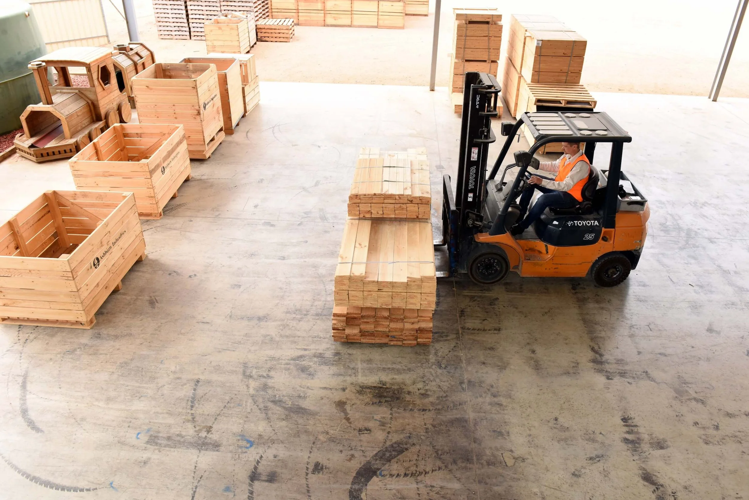 A worker operating a forklift on a concrete floor surrounded by stacked wooden crates and pallets in an indoor or covered outdoor space.