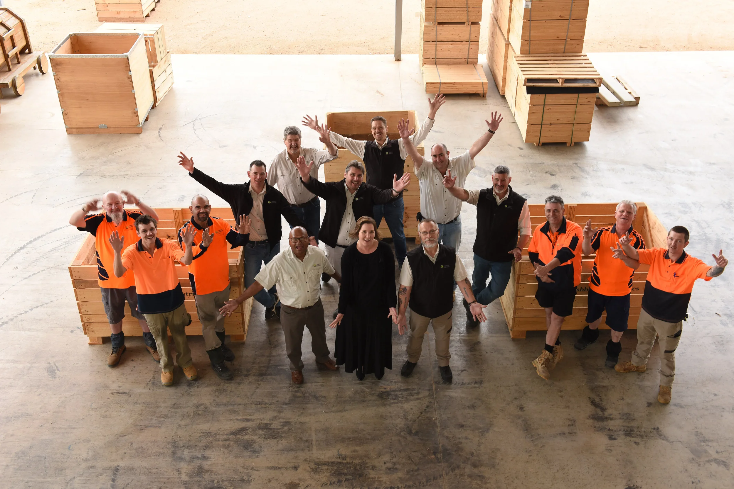 Group of workers and supervisors standing in a warehouse, smiling and cheering with arms raised, surrounded by stacks of wooden crates.