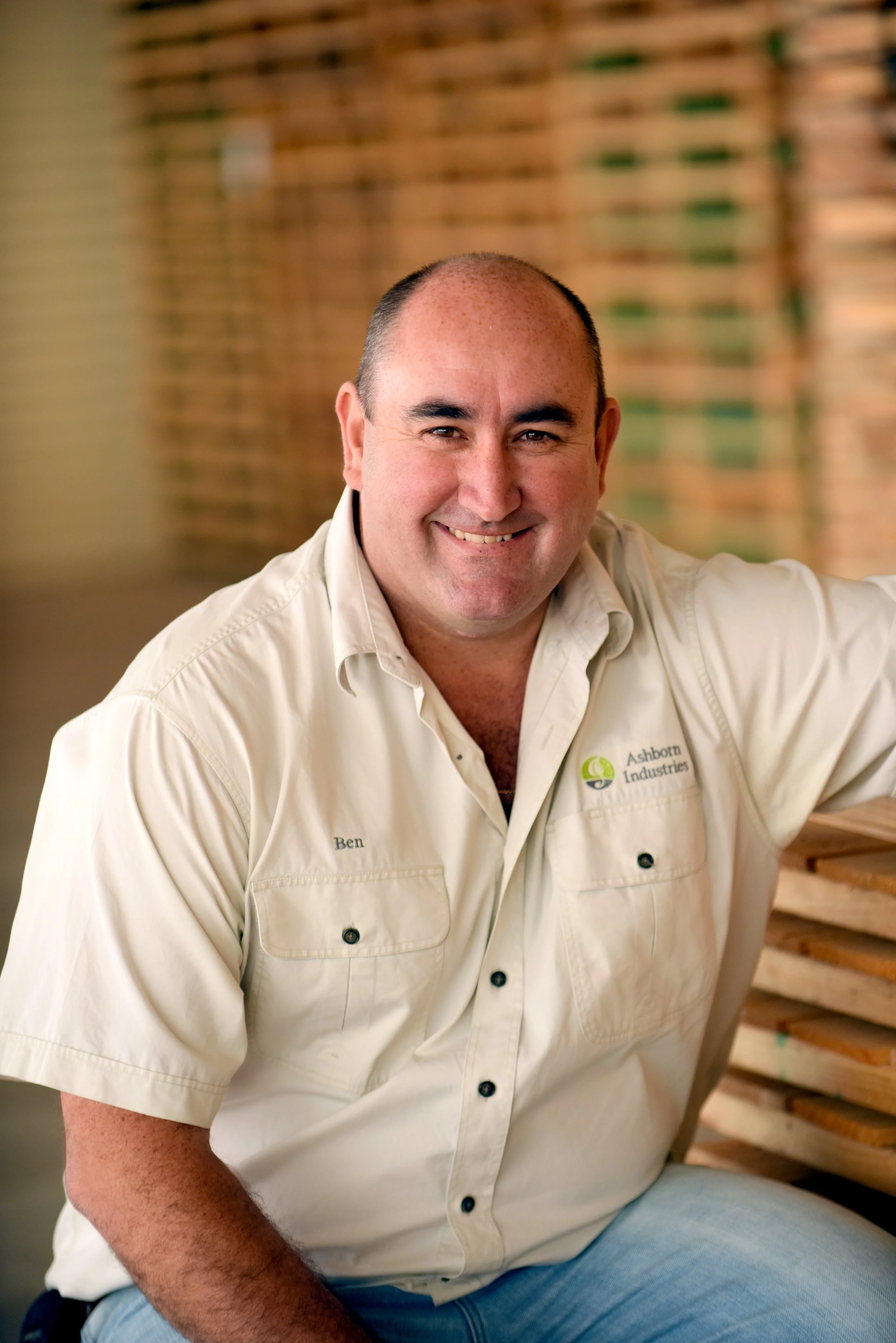 Stacks of wooden pallets inside a warehouse with a view of a farm field and trucks outside.