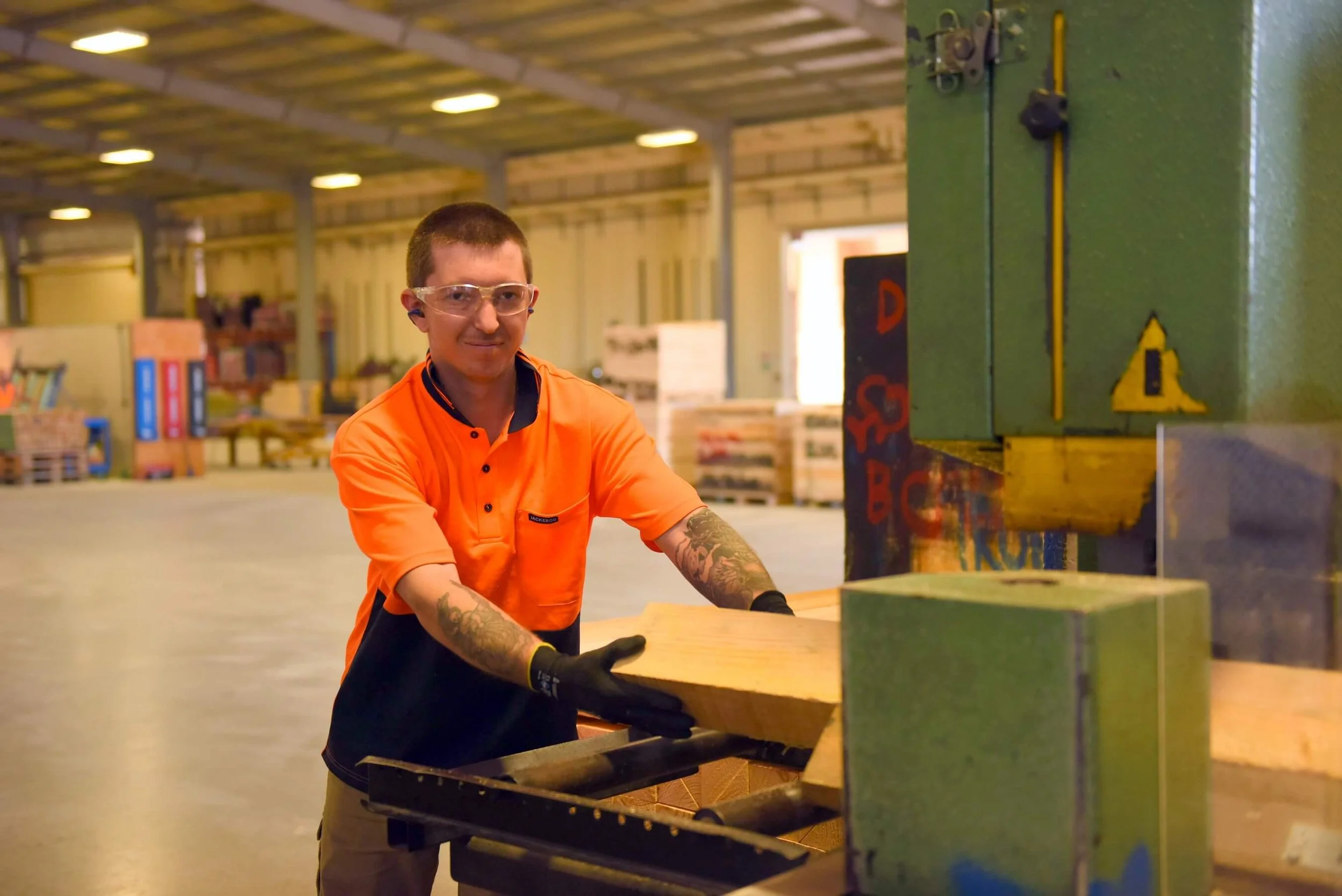 A man wearing safety glasses, ear plugs, and an orange work shirt operating a woodworking machine in a warehouse or lumber yard, holding a piece of wood.