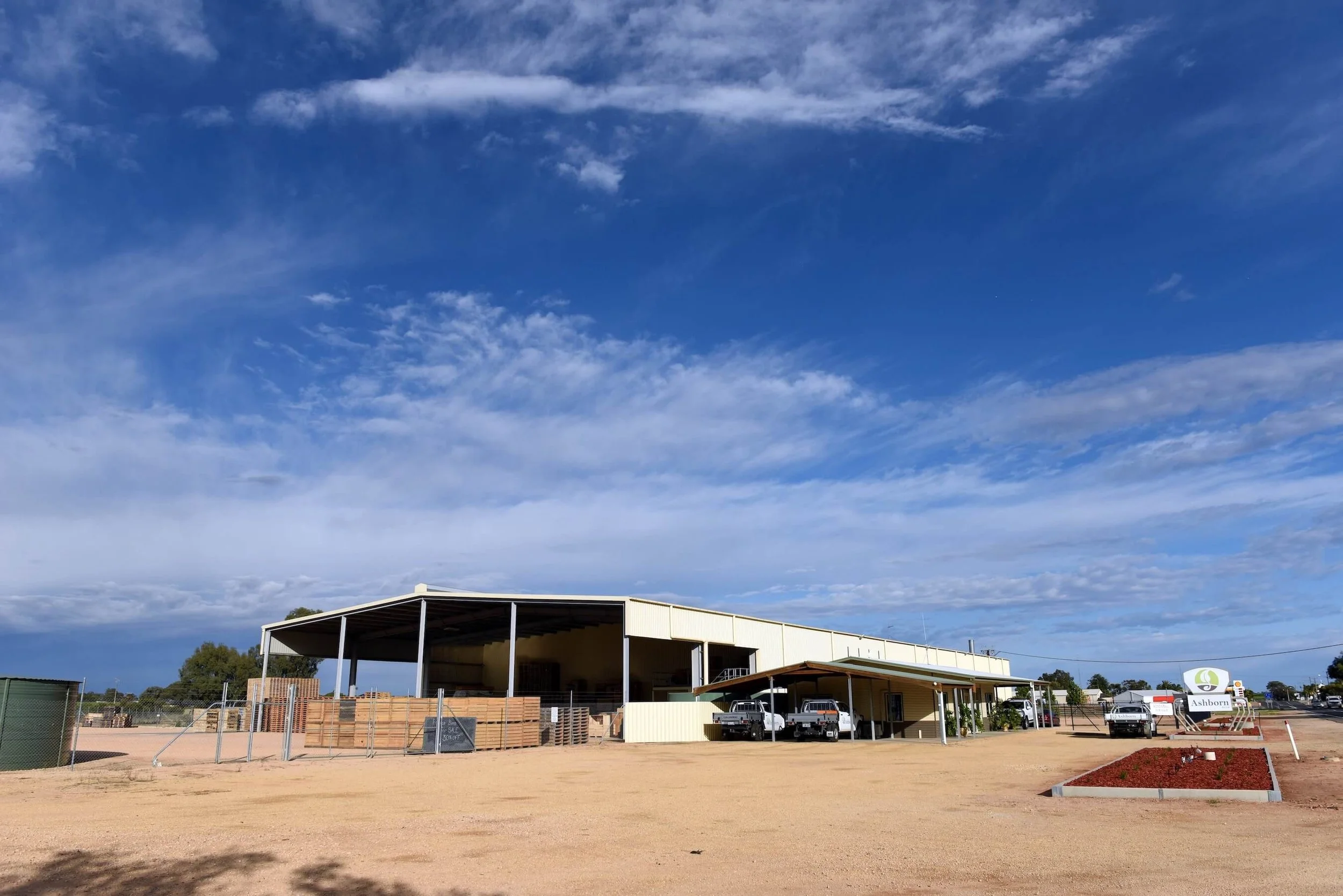 A large, open warehouse building on a dirt lot with a few trucks parked under its roof, a sign for Ashborn, and a flower bed in the foreground.