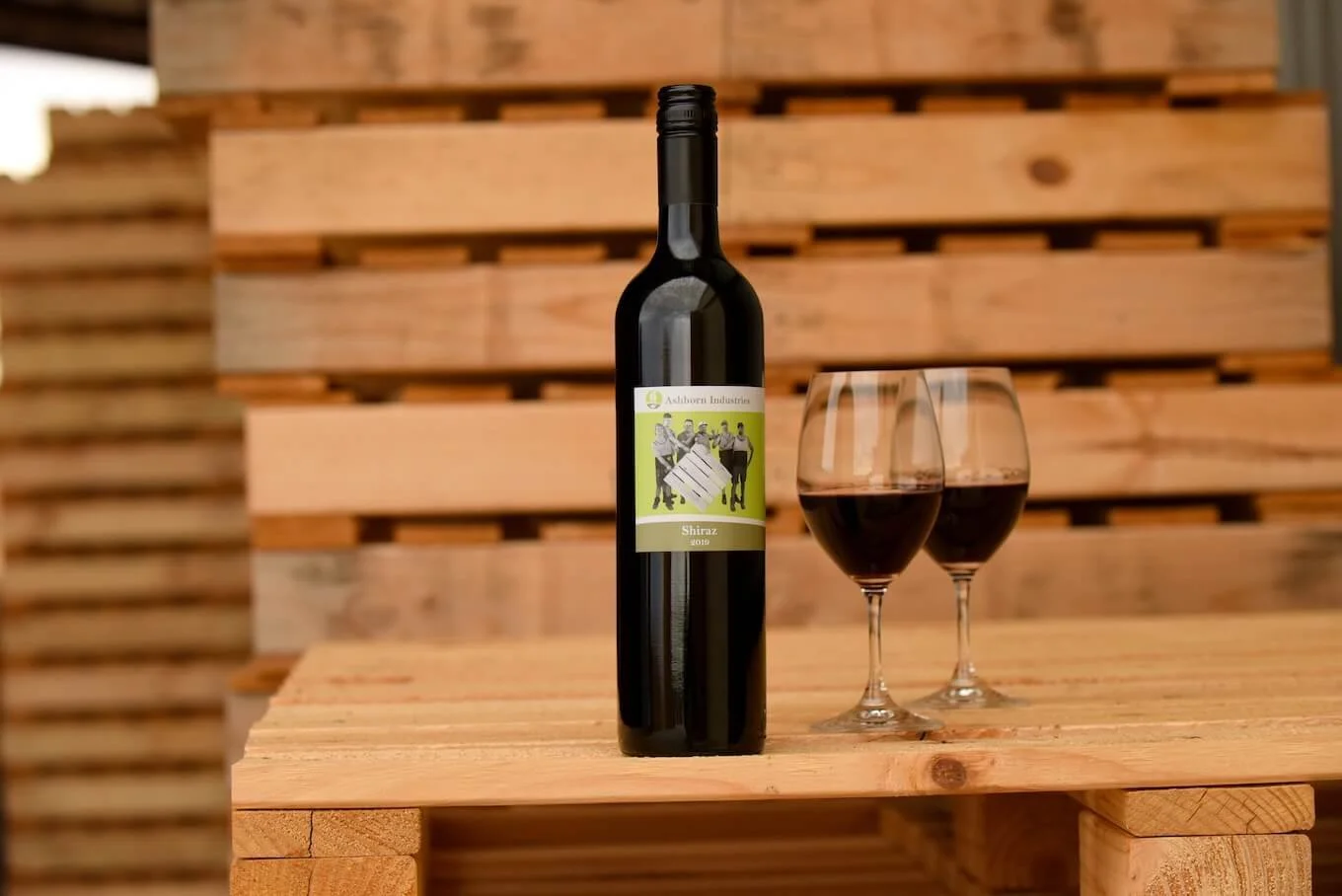 A bottle of red wine labeled 'Shiraz 2019' and two filled wine glasses are placed on a wooden table with a wooden pallet background.