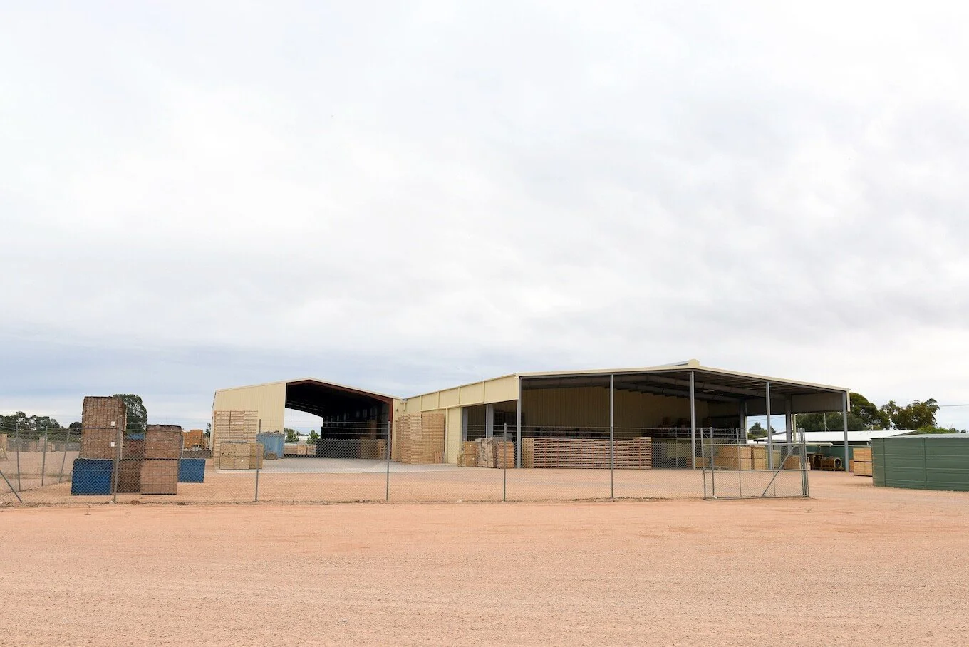 A large industrial warehouse or storage shed with an open front, surrounded by a chain-link fence, on a dirt lot with scattered pallets and crates, under a cloudy sky.