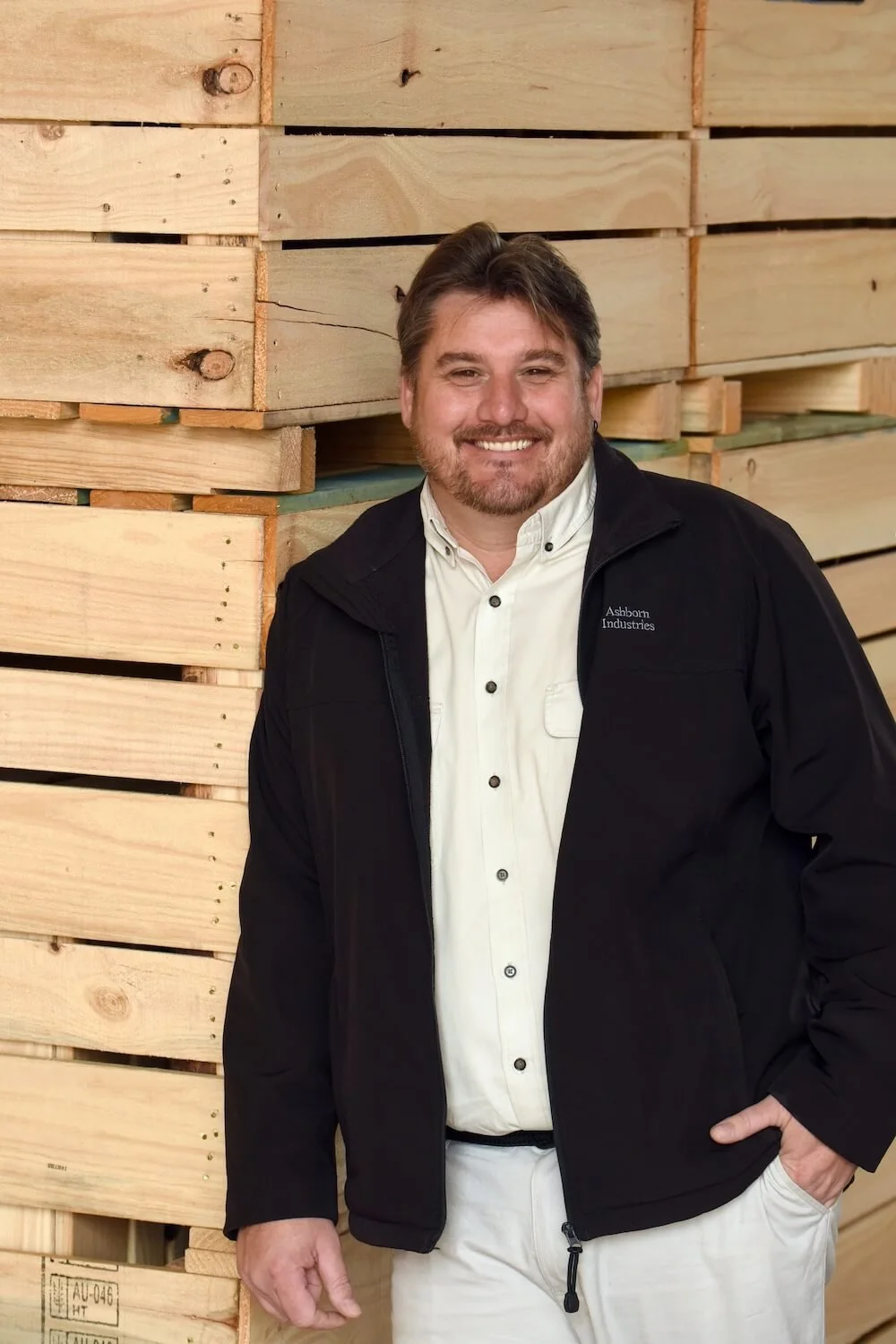 A man with brown hair, beard, and mustache smiling, wearing a white button-up shirt and black jacket, standing in front of a stack of wooden pallets.