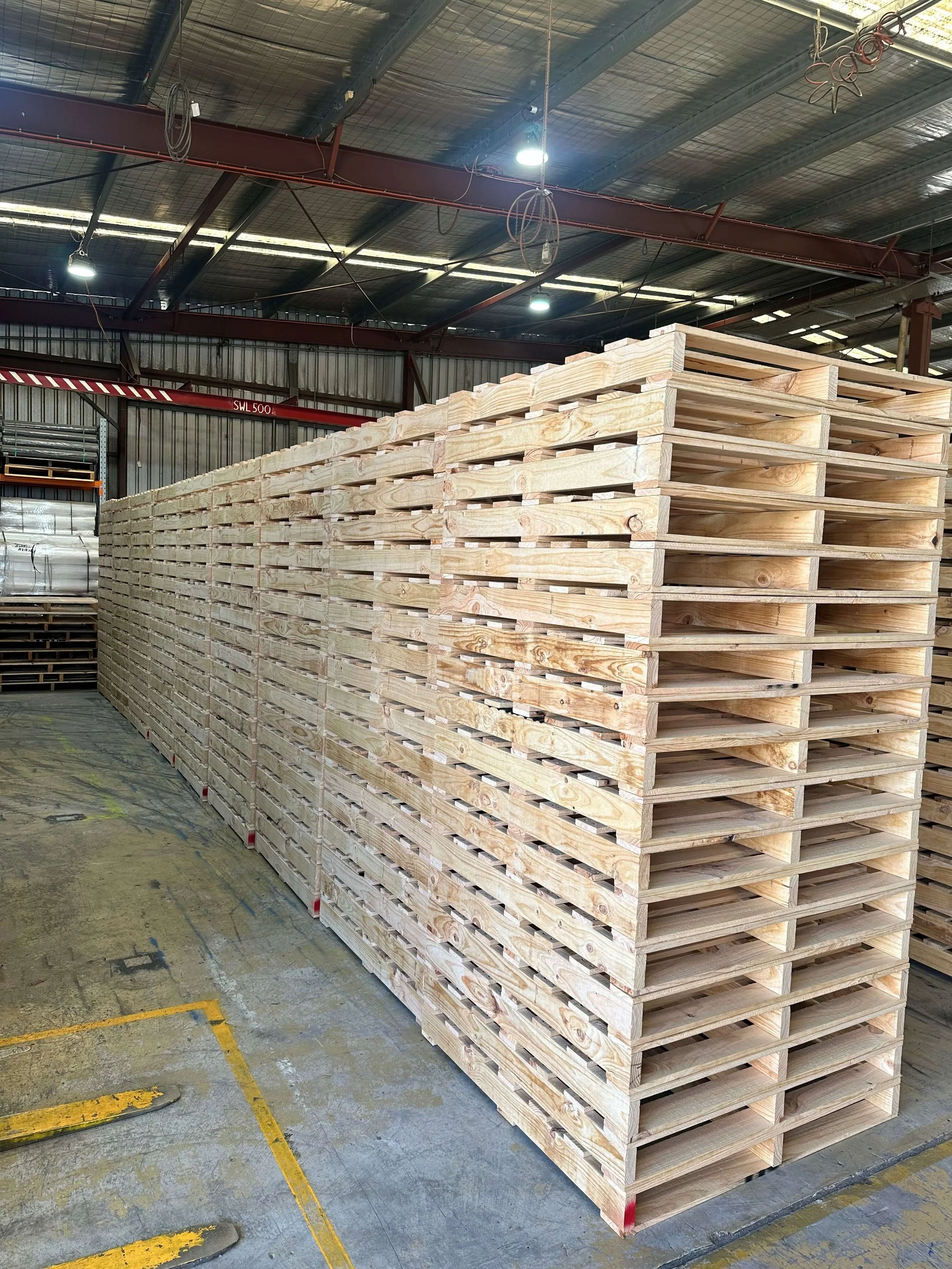 Stacked wooden pallets in an industrial warehouse with a metal roof, overhead lighting, and visible electrical wiring.