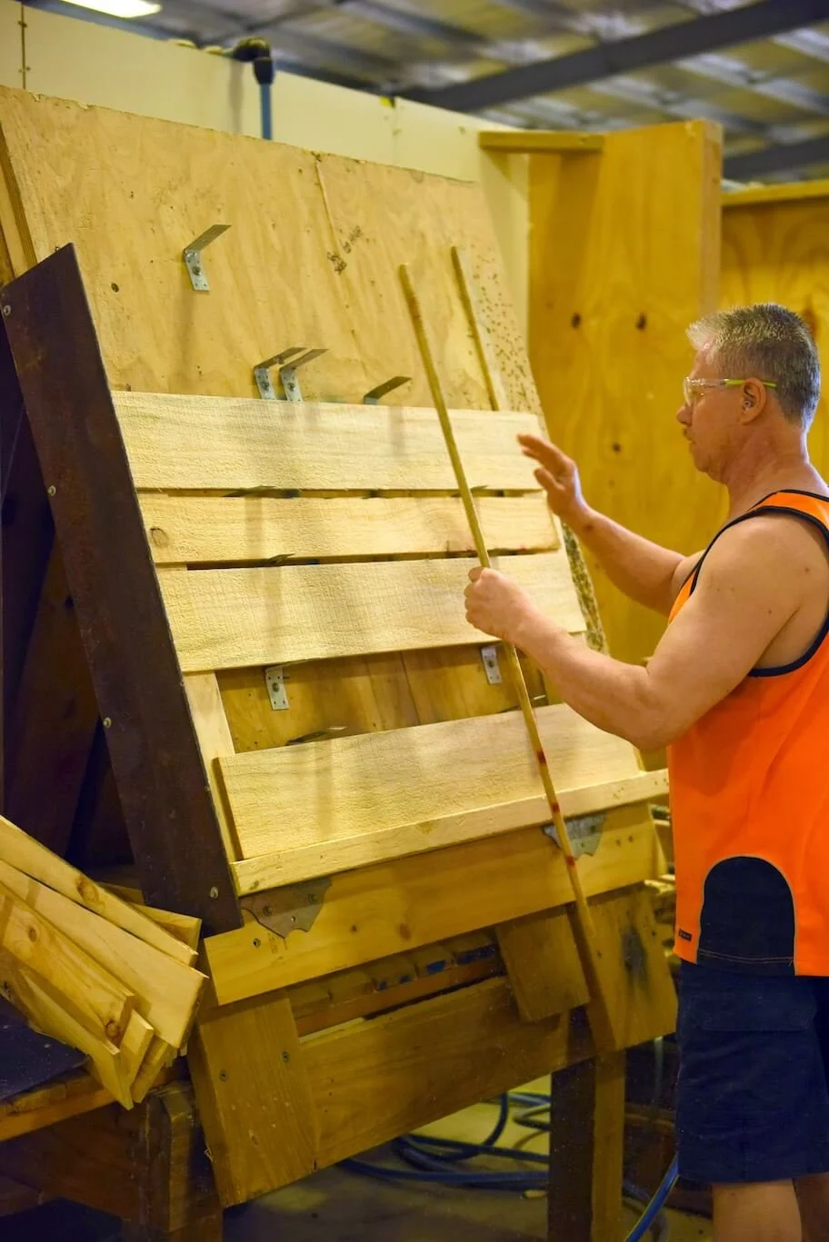 A man wearing safety glasses and an orange sleeveless shirt working on a woodworking project, sanding or finishing a wooden plank in a workshop.