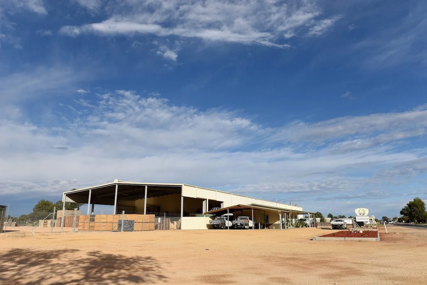 A large building with a metal roof and open sides, situated in a sandy area with a few cars parked nearby. There is a sign that reads "Ashbourne" and a landscaped brick planter in front of it under a partly cloudy sky.
