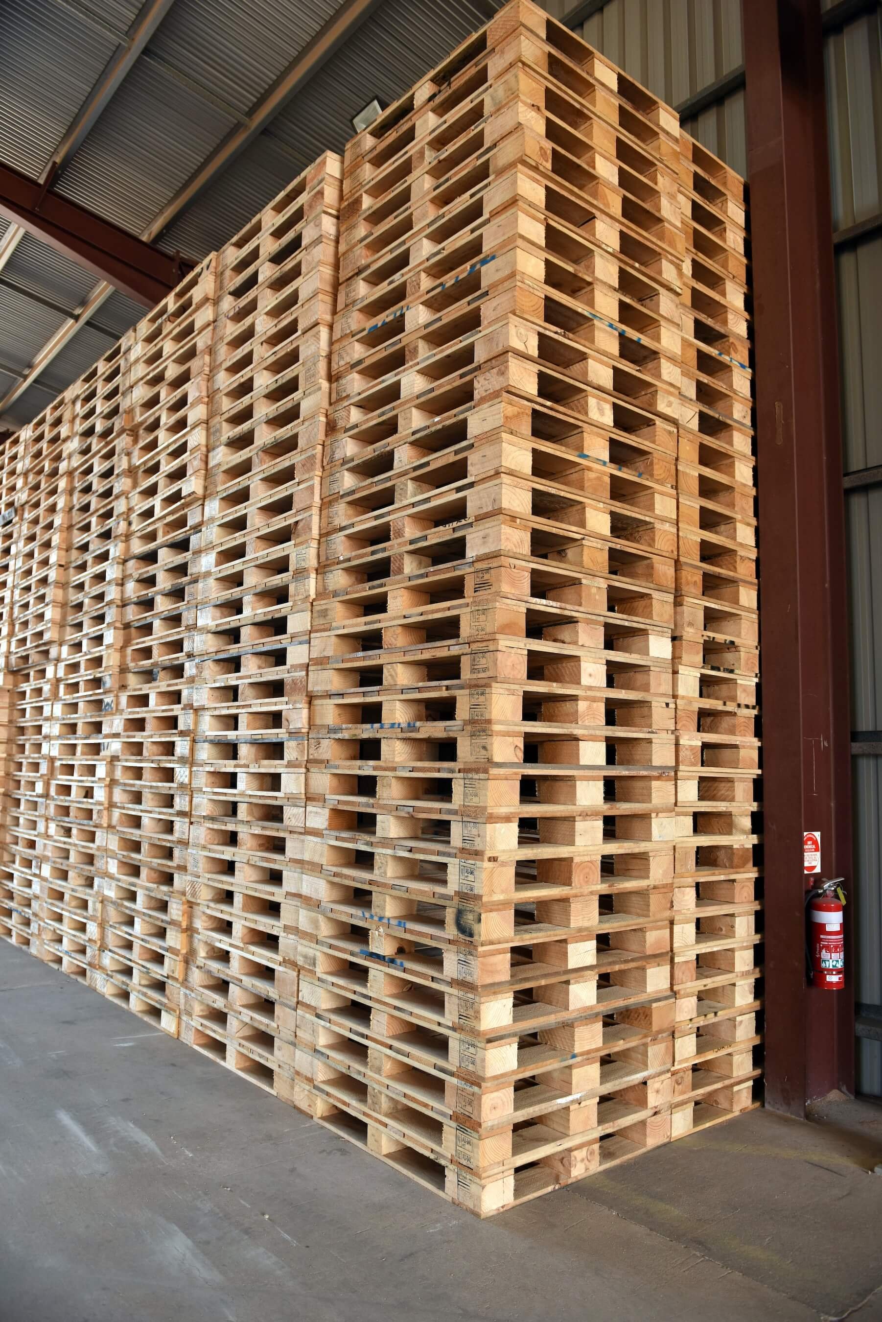 Large stack of wooden pallets inside a warehouse with metal walls and roof, and a fire extinguisher mounted on the wall to the right.