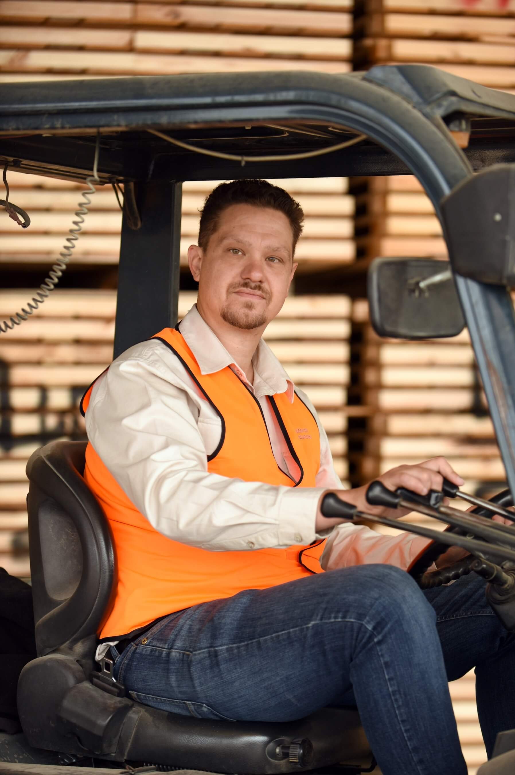 A man sitting on a forklift, wearing a white shirt and an orange safety vest, indoors with stacks of wood in the background.