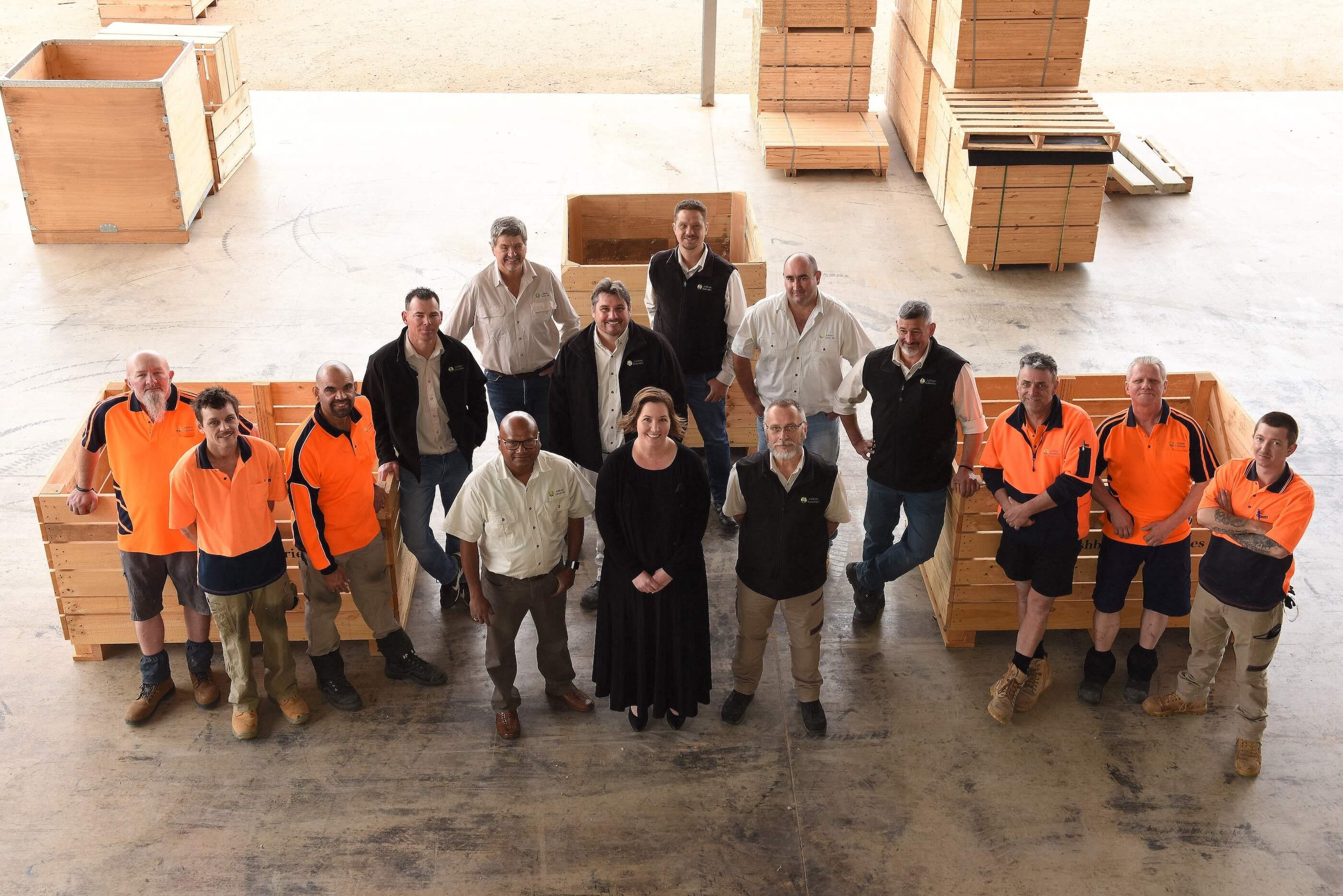 A group of fifteen people standing inside a warehouse or factory with wooden crates around them. Five people are wearing orange work uniforms, while the others are in casual or business attire. They are all looking up at the camera.