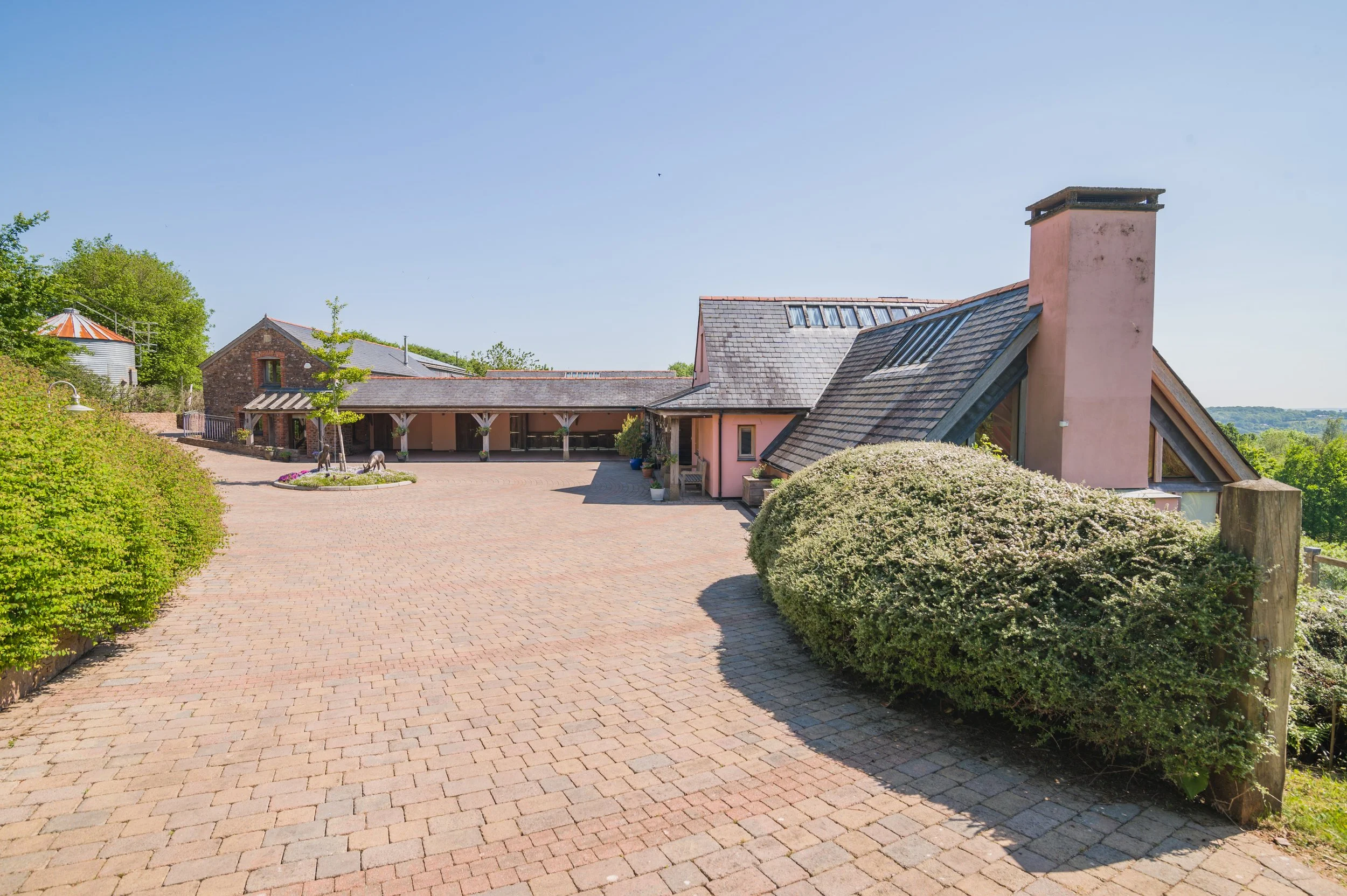 A sunny day view of a house with a brick driveway, surrounded by greenery, with a chimney, windows, and a pink exterior.