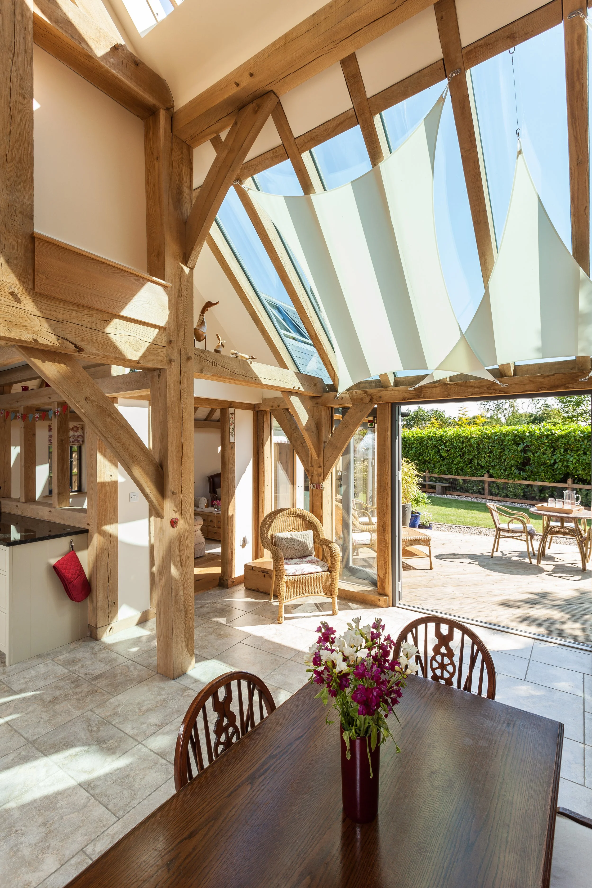 Sunlit interior of a house with exposed oak beams, a wooden dining table with a flower vase, wicker chairs, and large glass doors opening to a backyard patio with outdoor furniture and greenery.
