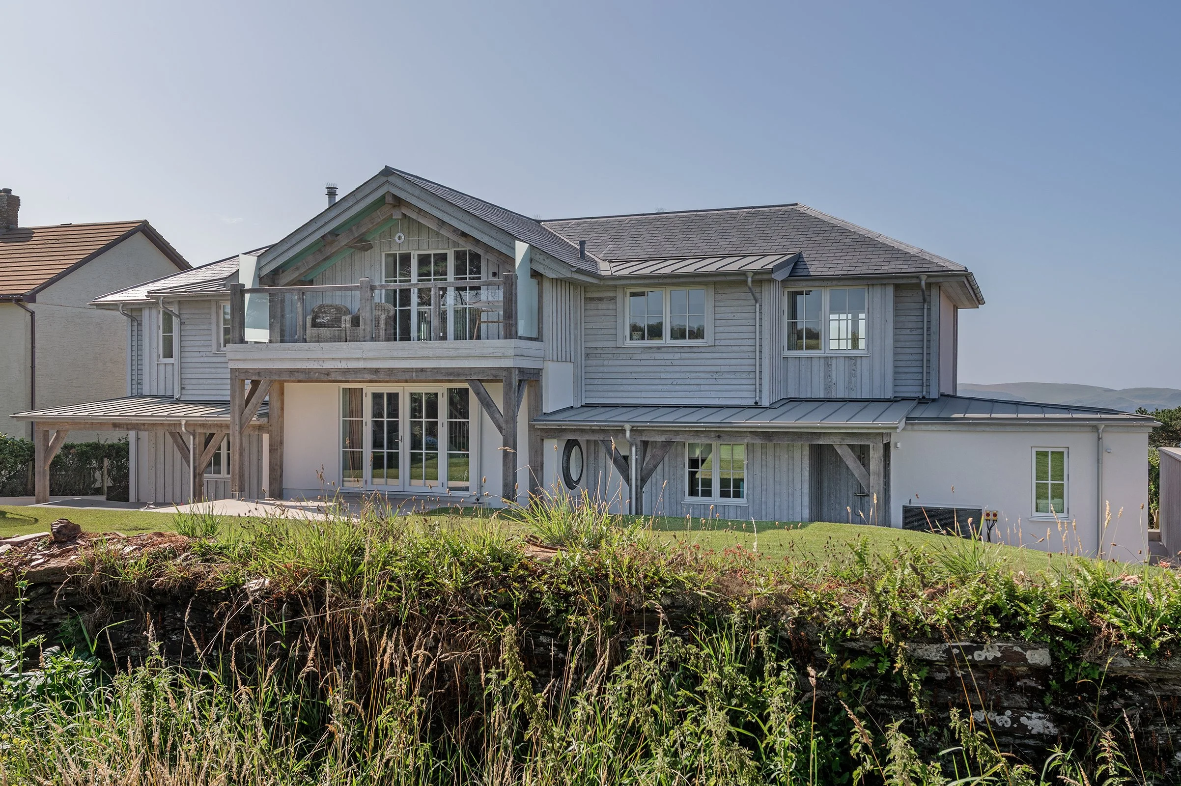 A two-story timber framed house with a combination of wood and white exterior siding, large windows, a balcony on the upper level, and a landscaped front yard with grass and plants.