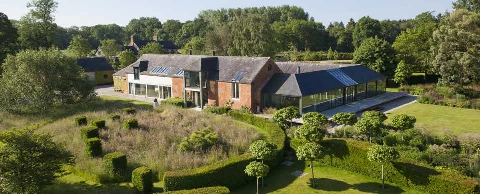 A large modern house with brick and glass exterior, surrounded by landscaped gardens with trimmed bushes and trees, and open grassy areas.