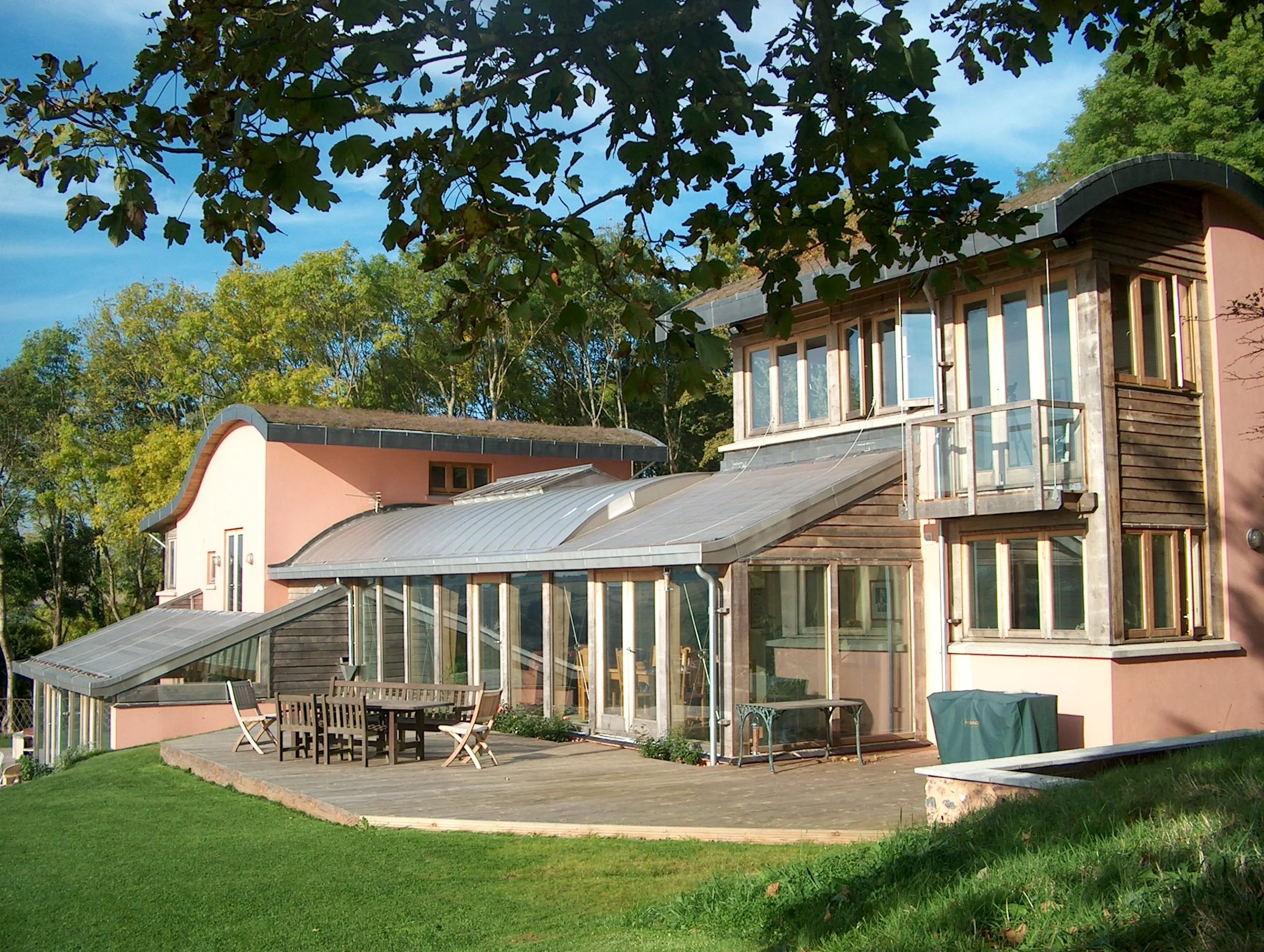 A modern house with pink and timber exterior, multiple large windows, a wooden deck with outdoor dining furniture, and surrounded by green grass and trees.