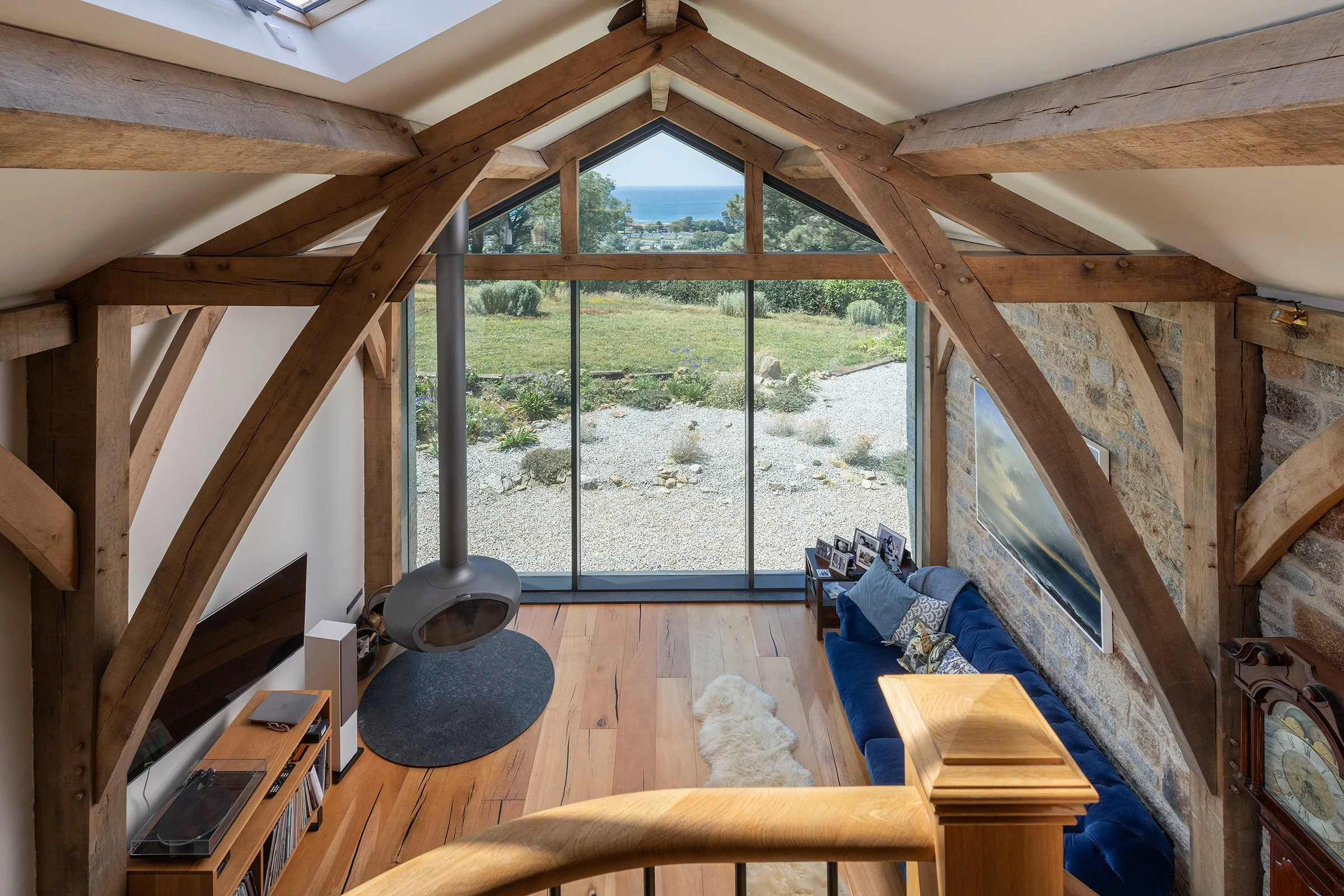 A living room with large triangular floor-to-ceiling windows overlooking a garden and ocean view. Inside features exposed wooden beams, a stone wall, a blue sofa with pillows, a small wooden coffee table, a sheepskin rug, and a modern wood stove with a metal pipe.