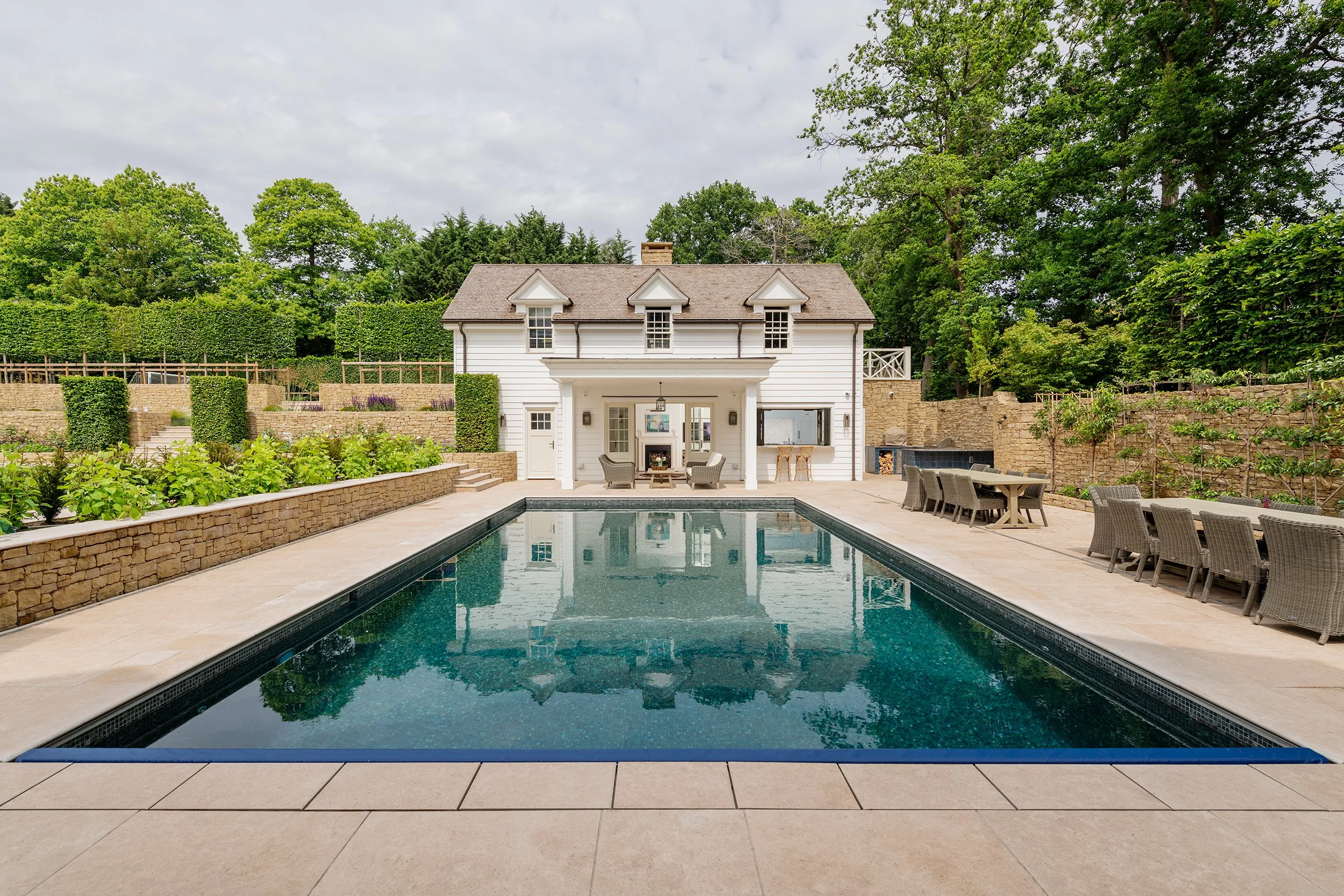 Swimming pool, white house, outdoor furniture, and lush green trees in the background.