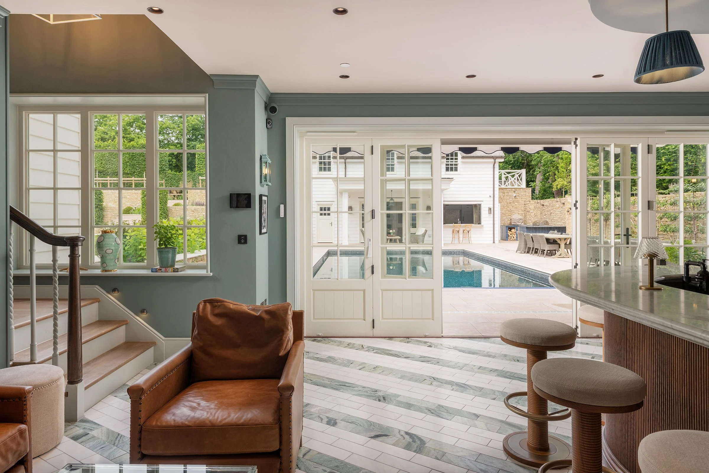 Living room with large windows and open sliding glass doors leading to a pool area. There are leather chairs, a bar with stools, and a staircase.