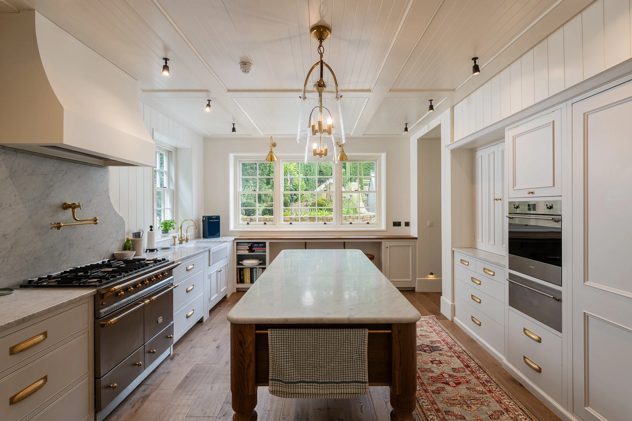 Bright modern kitchen with white cabinets, a large island with a marble top, gold hardware, stainless steel appliances, and large windows overlooking greenery.