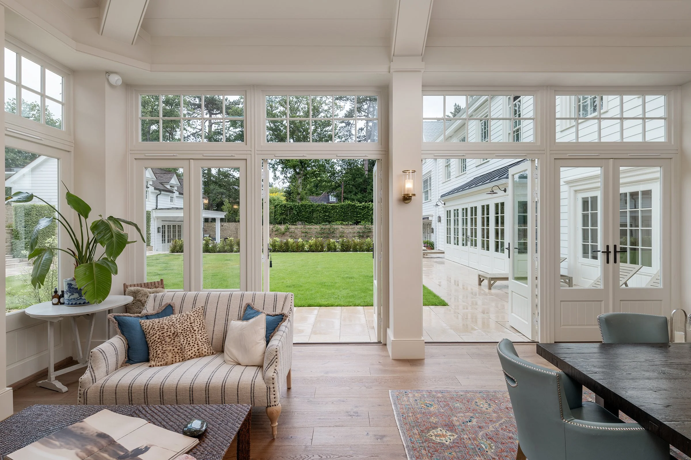 Interior view of a sunroom with large windows and a sliding glass door opening to a lawn and garden. There is a striped armchair with pillows, a side table with a large plant, and part of a dark wood dining table with light-colored chairs visible.