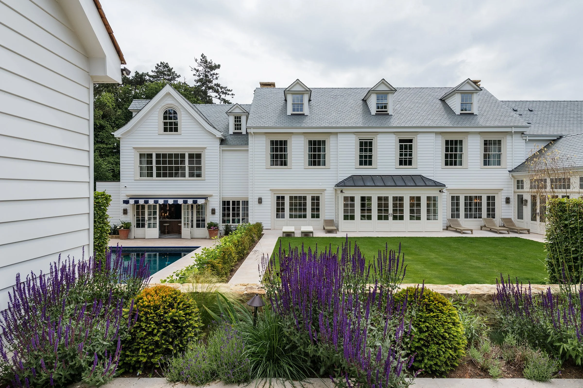 Backyard of a white house with large windows, a swimming pool, a lawn, and landscaped garden with purple flowers and bushes.