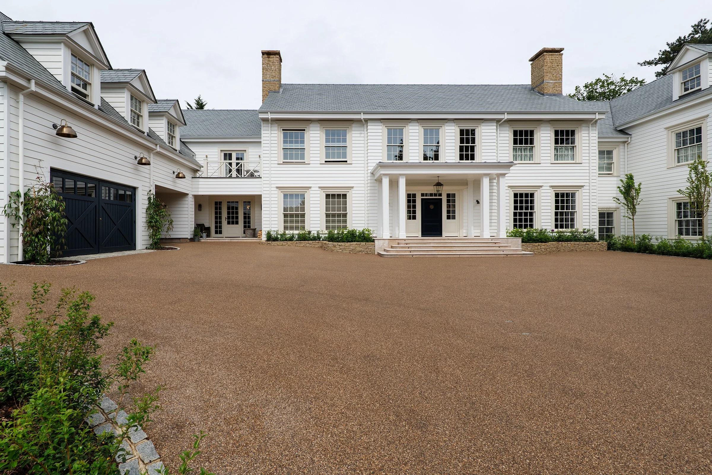 Large white house with multiple windows, black front door, and a grand staircase leading to the entrance. There is a gravel driveway and some small plants and shrubs in front. The house has a gray roof and brick chimneys.