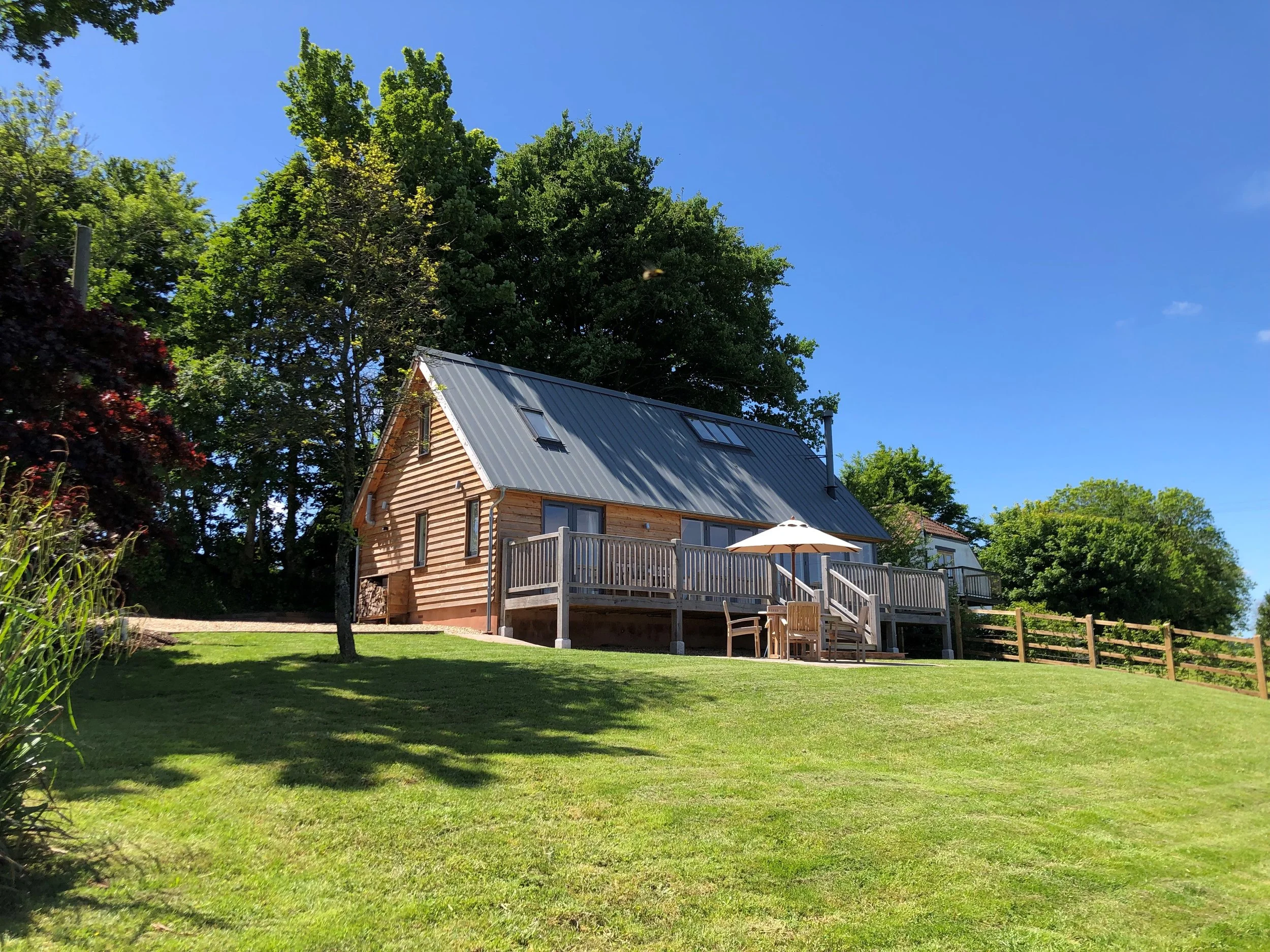 A wooden house with a metal roof, balcony, and outdoor furniture under a white umbrella on a grassy lawn, surrounded by trees under a clear blue sky.