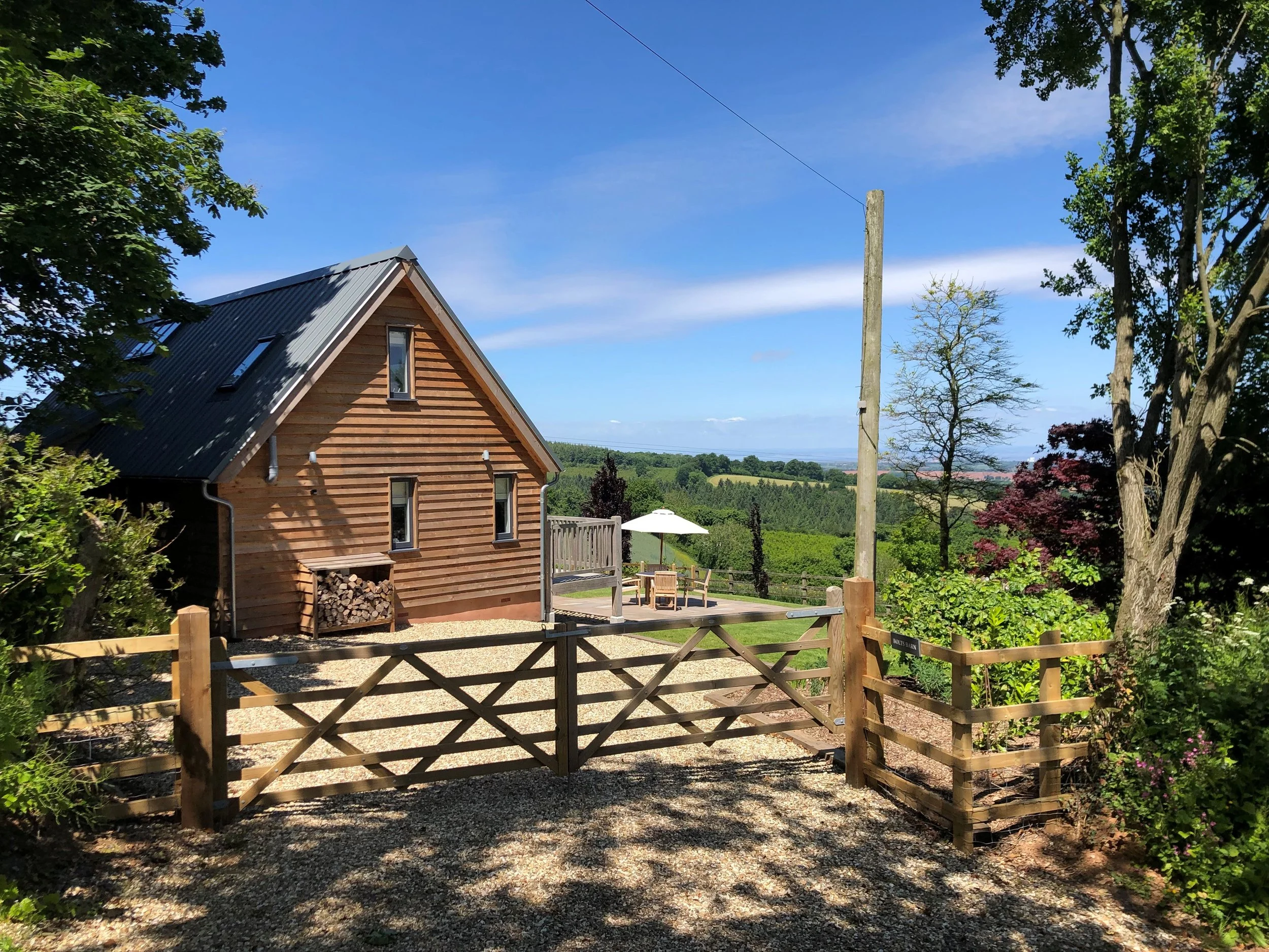A wooden house with a gray metal roof, surrounded by trees, with a gravel driveway and a garden area featuring outdoor furniture and an umbrella, overlooking a scenic landscape of rolling hills and green fields under a blue sky.