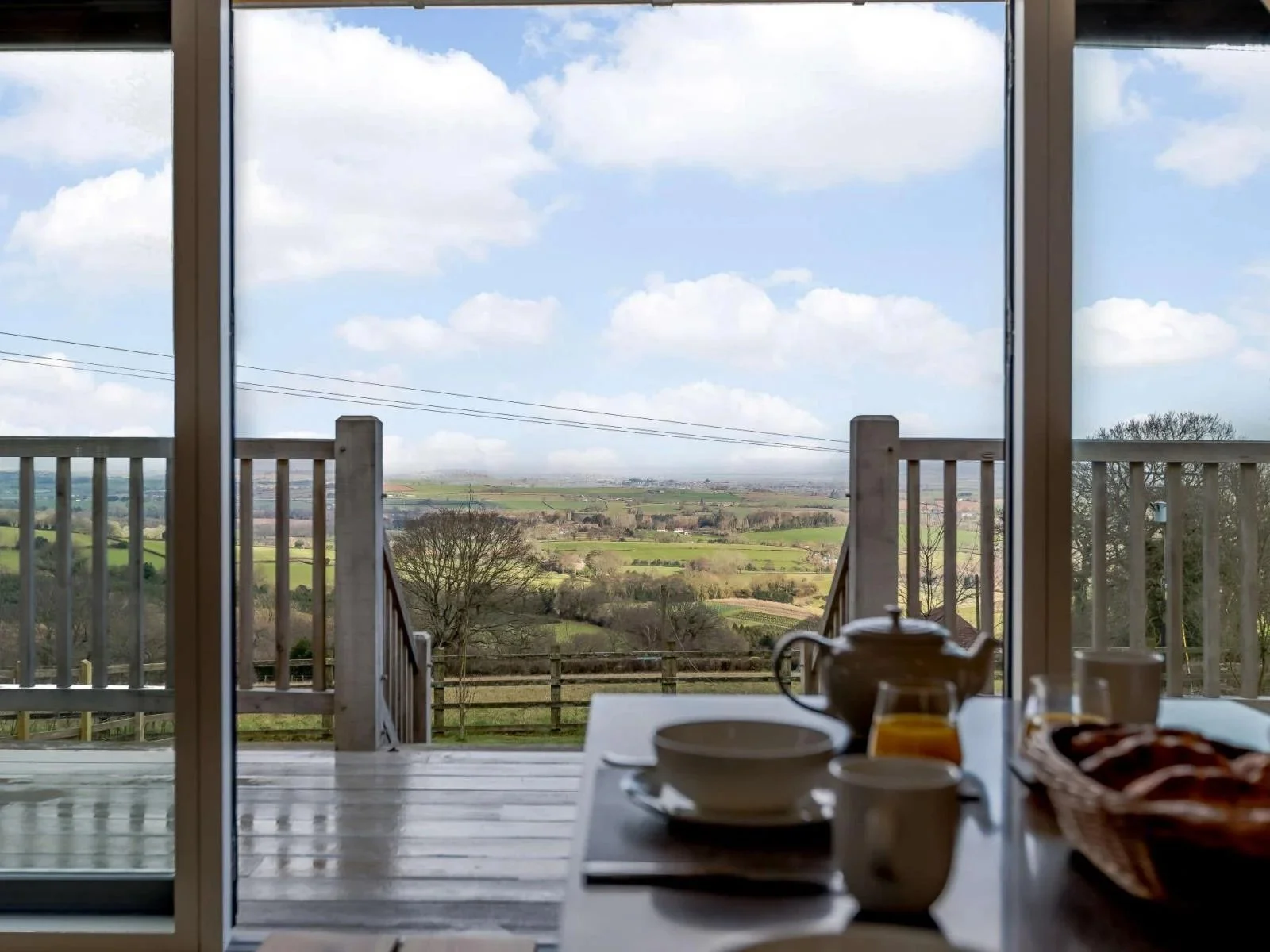 A dining table with a teapot, cups, and a basket of bread, looking out through a large sliding glass door onto a backyard with a wooden deck and a view of rolling green hills and trees under a partly cloudy sky.
