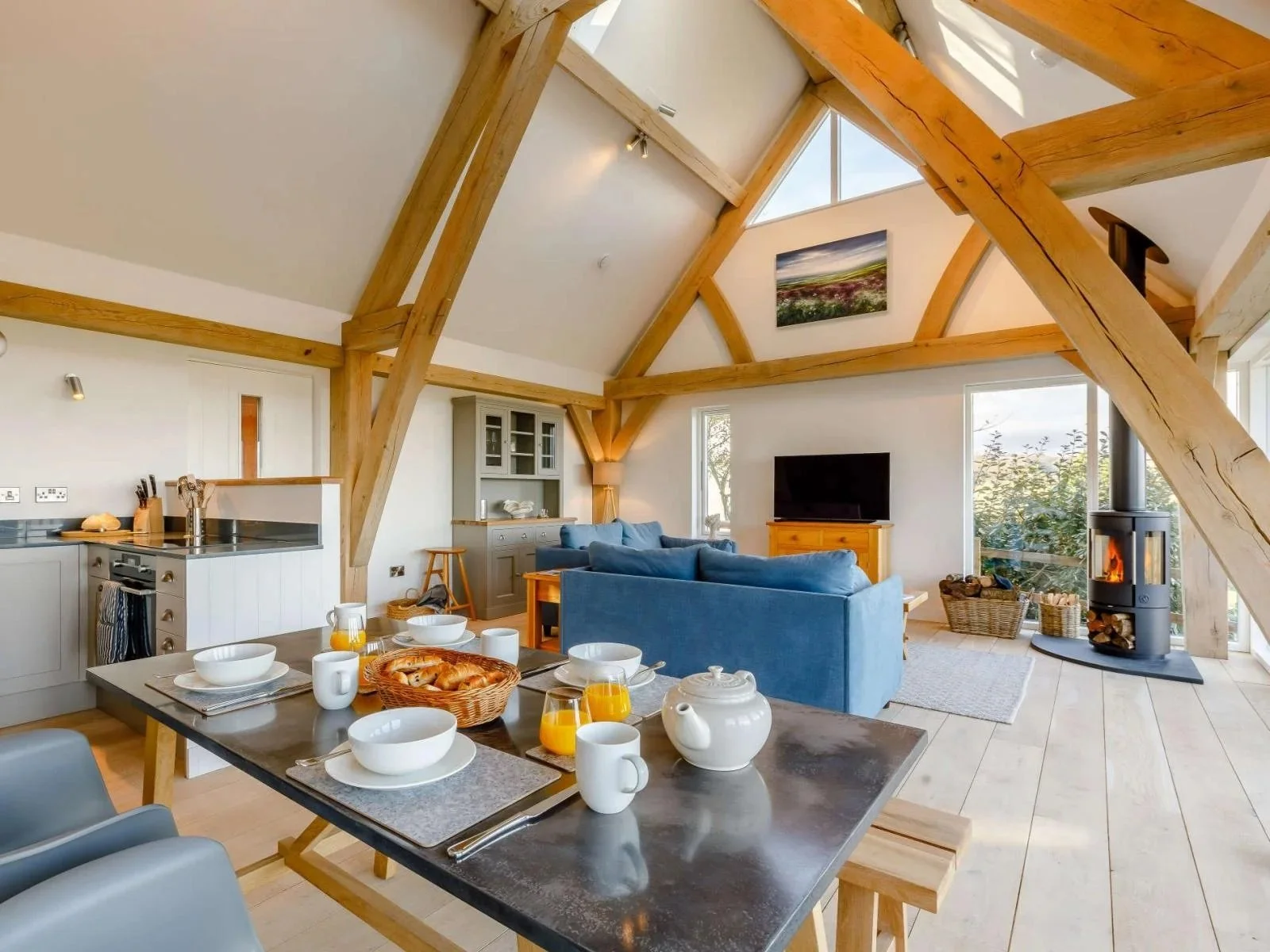 Open-concept living and dining area in a cozy home with wooden beams, a blue sofa, a fireplace, a dining table set with breakfast, a television, and large windows with natural light.