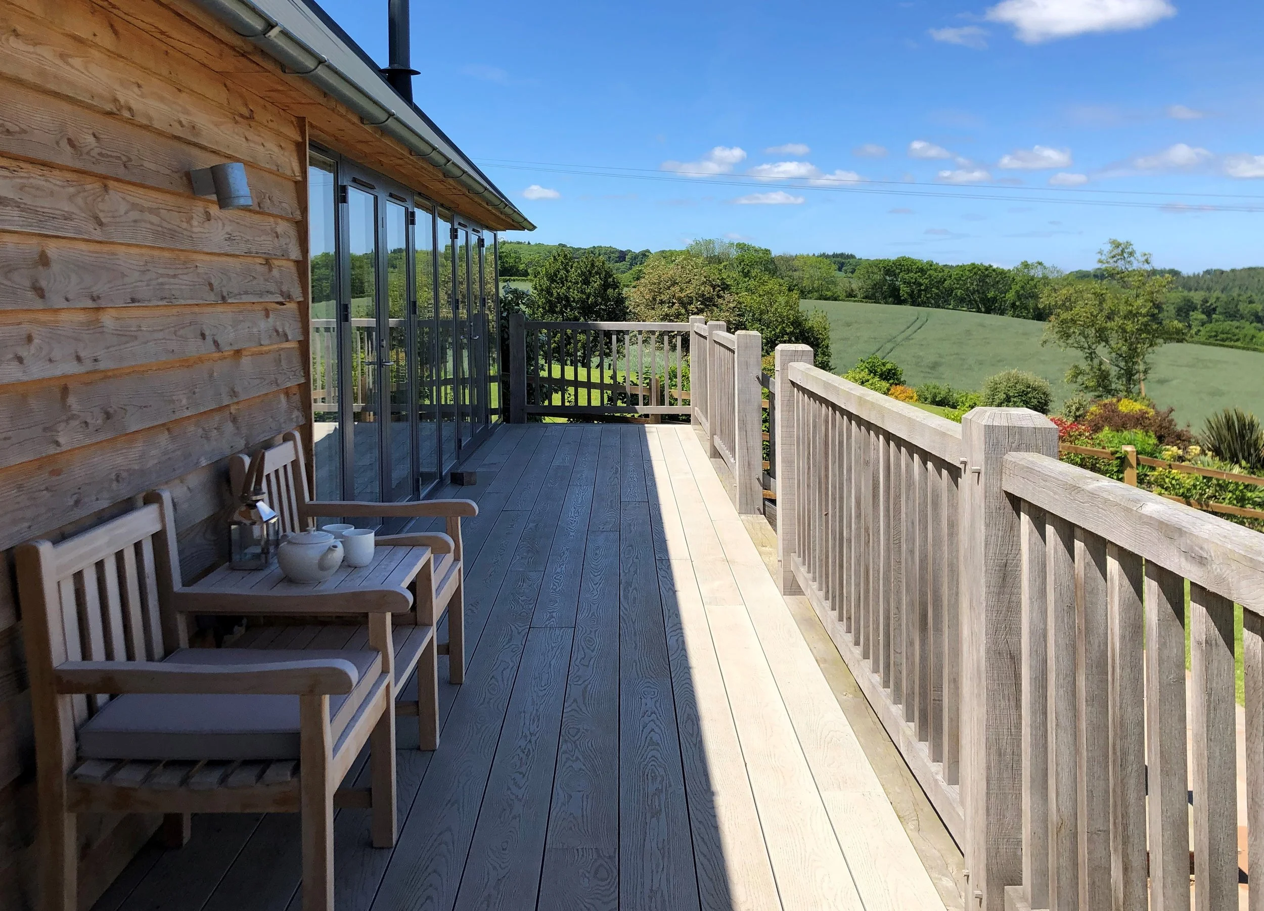 A wooden deck attached to a house with sliding glass doors, overlooking a lush green landscape with trees and rolling hills under a partly cloudy blue sky.