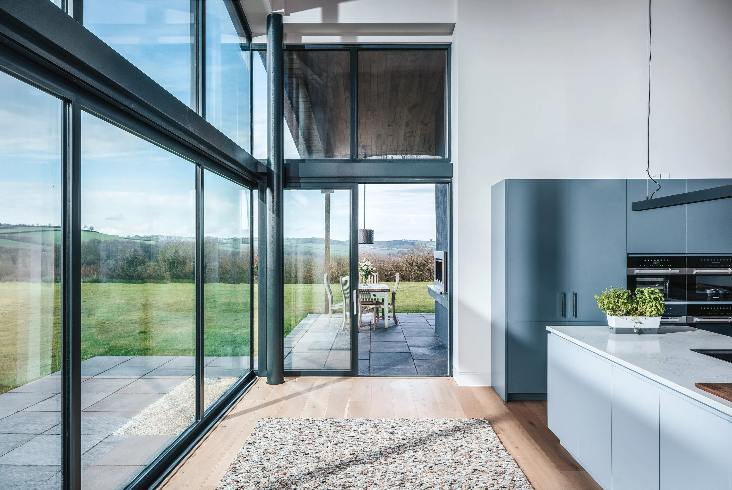 Modern kitchen with large floor-to-ceiling glass windows showing an outdoor patio and green landscape, with a dining table and chairs visible outside.