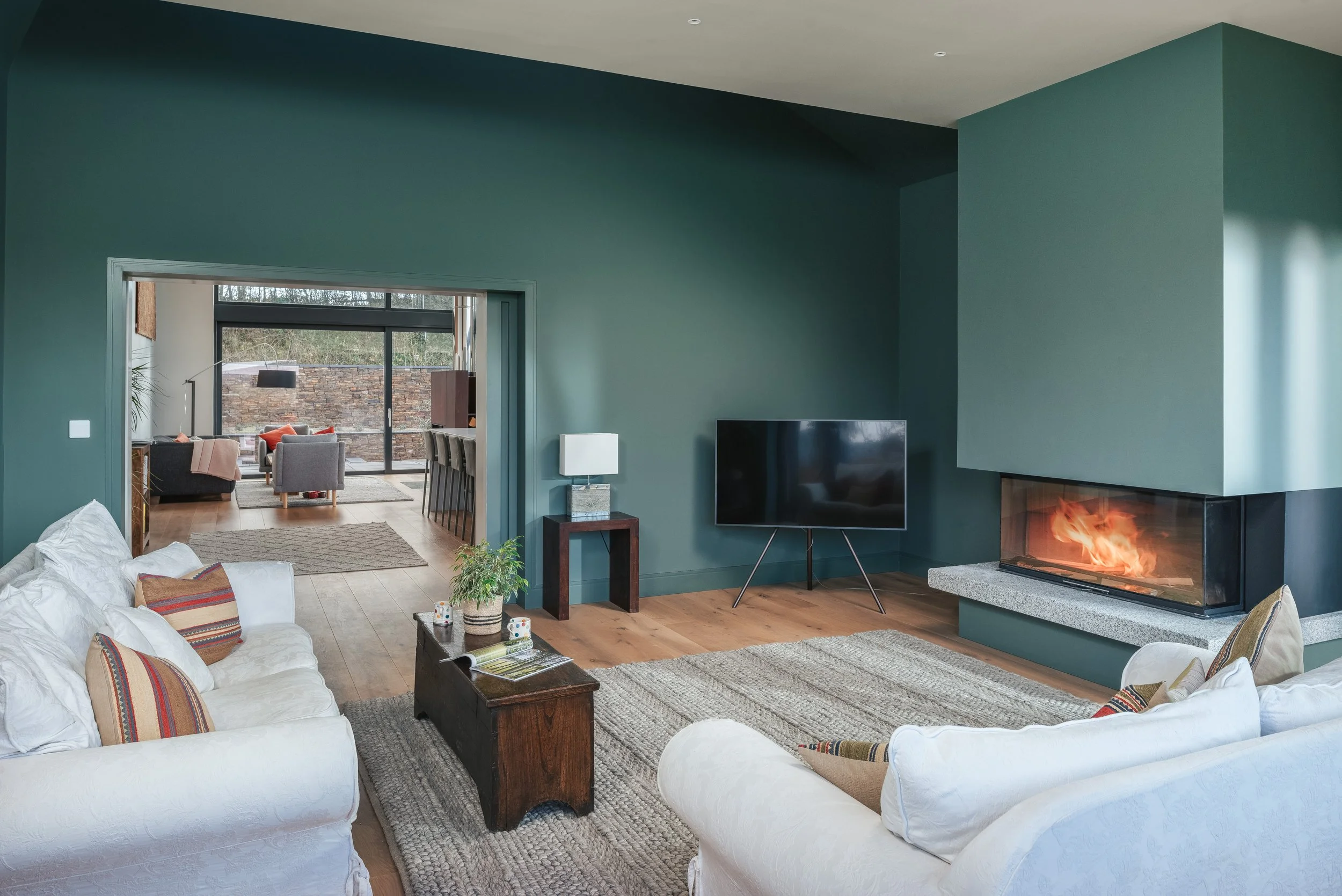 Living room with green walls, white sofas with colorful striped pillows, a wooden coffee table, a gray rug, a flat-screen TV, and a modern fireplace with fire. An open doorway leads to a dining area with a gray seating area and large glass doors.