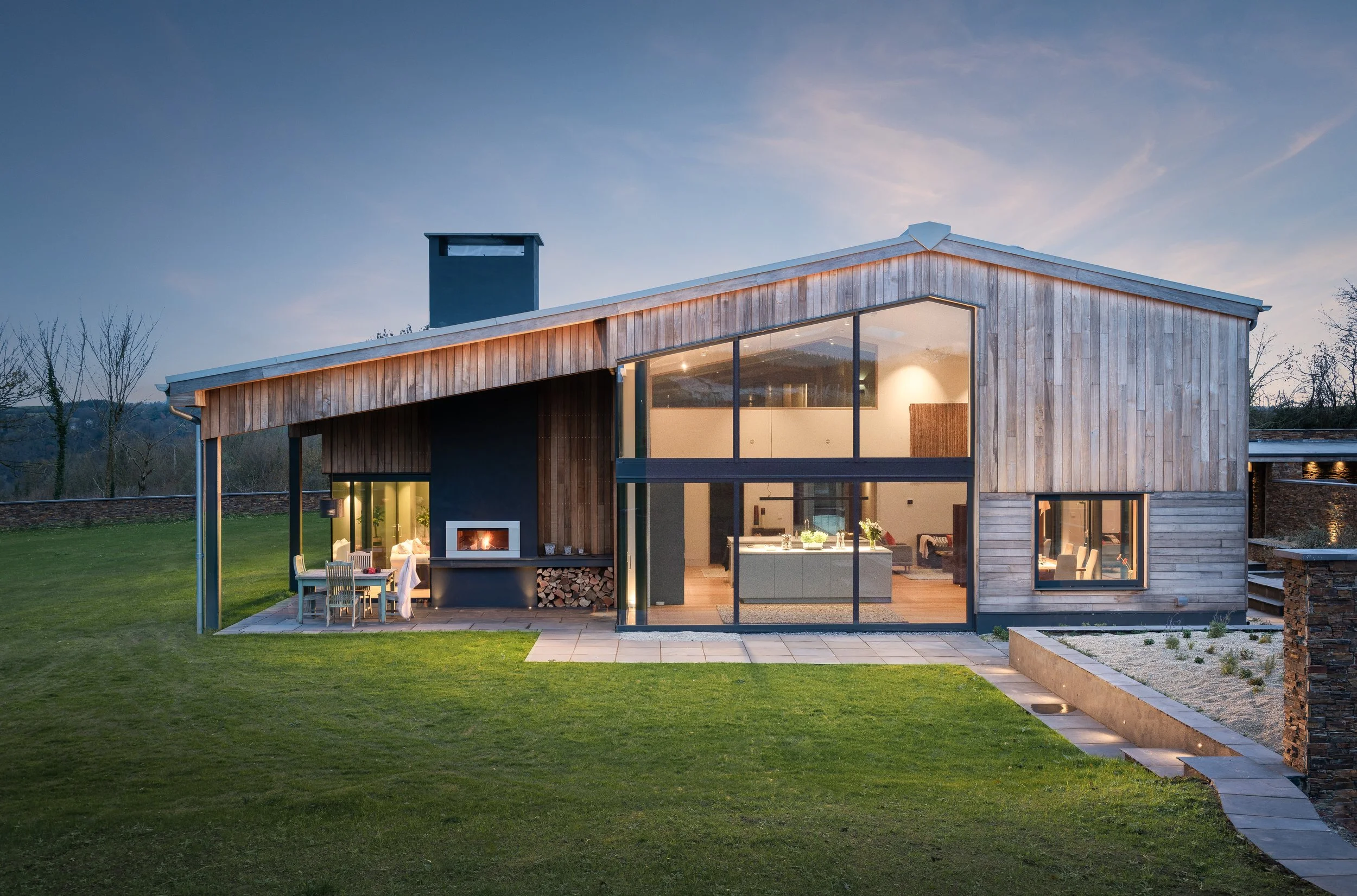 Modern two-storey house with large glass windows, wooden exterior, outdoor dining area, fireplace, and neatly landscaped yard at dusk.