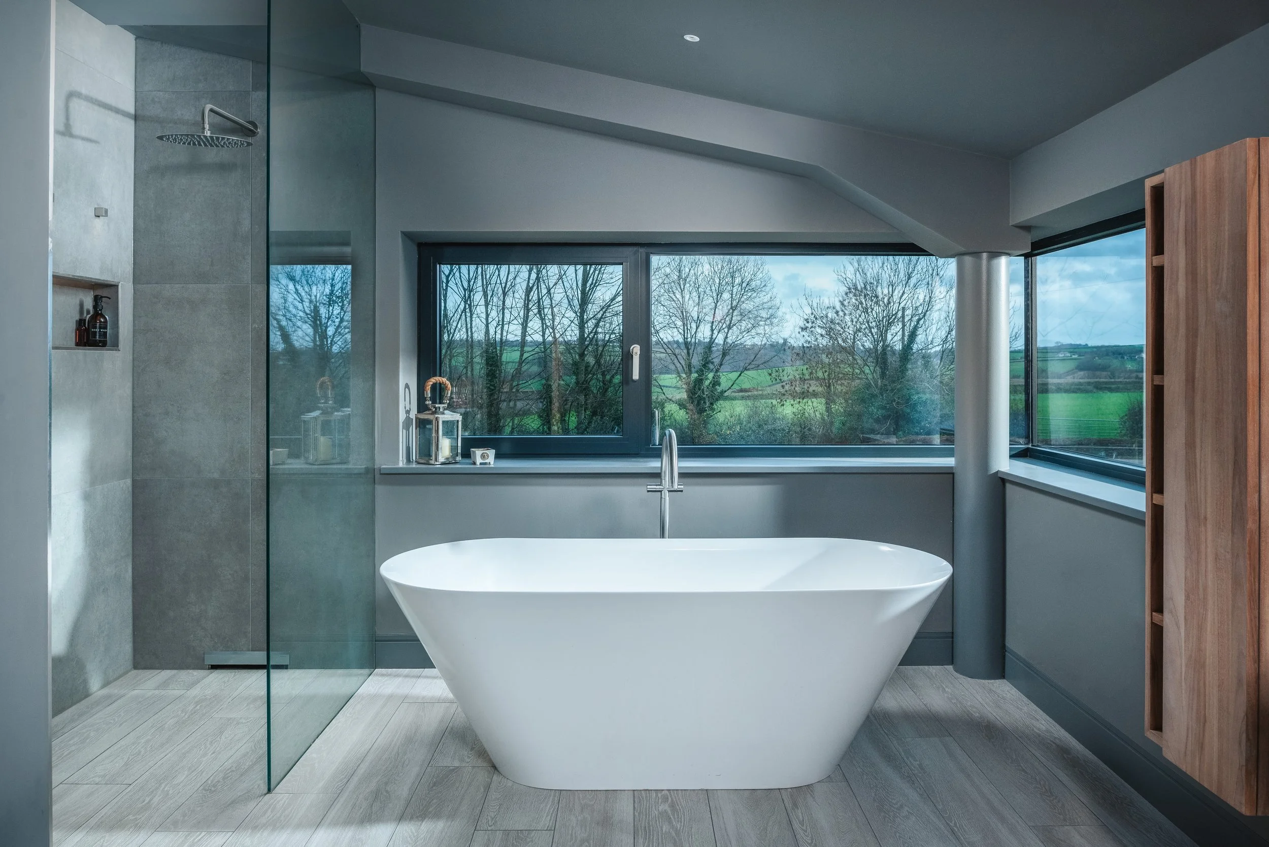 Modern bathroom with a freestanding white bath, large window with a view of trees and fields, wooden cabinet on the right, and a glass-enclosed shower on the left.