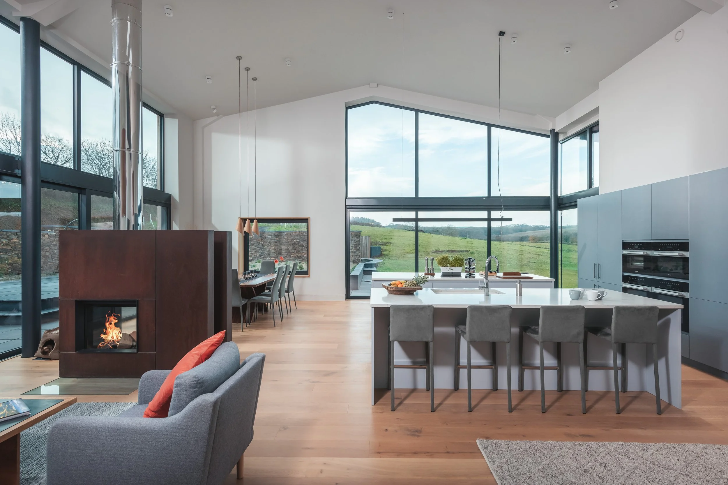 Modern open-concept living space with large windows, a kitchen island with bar stools, a fireplace, and a dining area, overlooking a green landscape.
