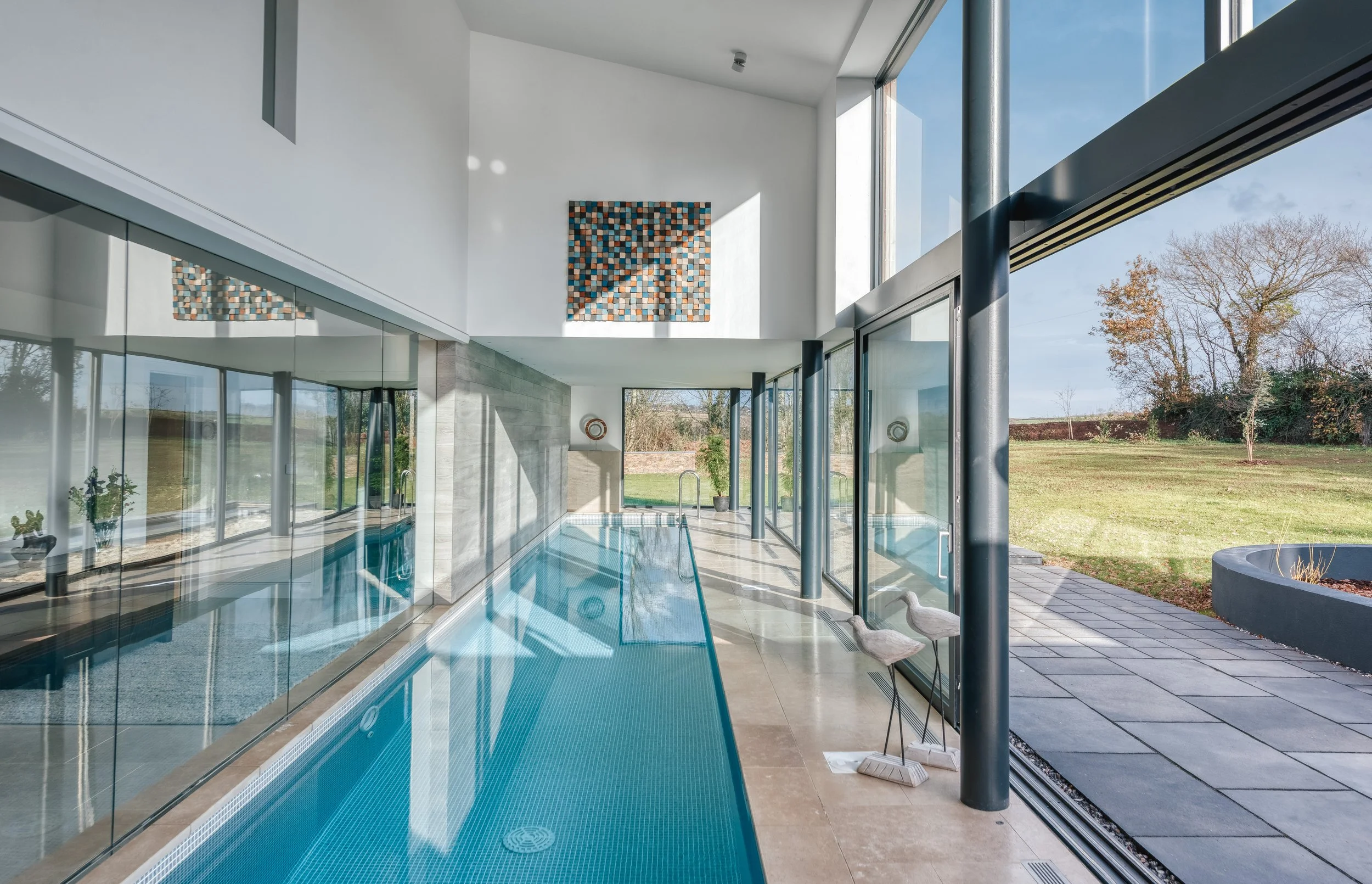 Modern indoor swimming pool with large glass windows and an outdoor garden view, featuring contemporary decor and sculptures of birds.