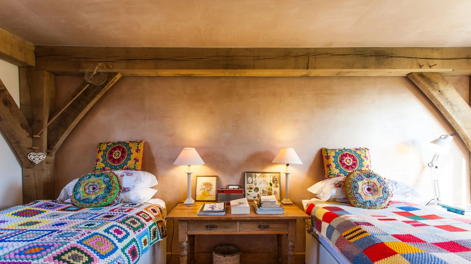 A cozy bedroom with two beds covered in colorful crochet blankets, and decorative pillows. A wooden nightstand between the beds holds lamps, picture frames, and books. The wall is warmly lit with soft lighting, and the room features exposed wooden be